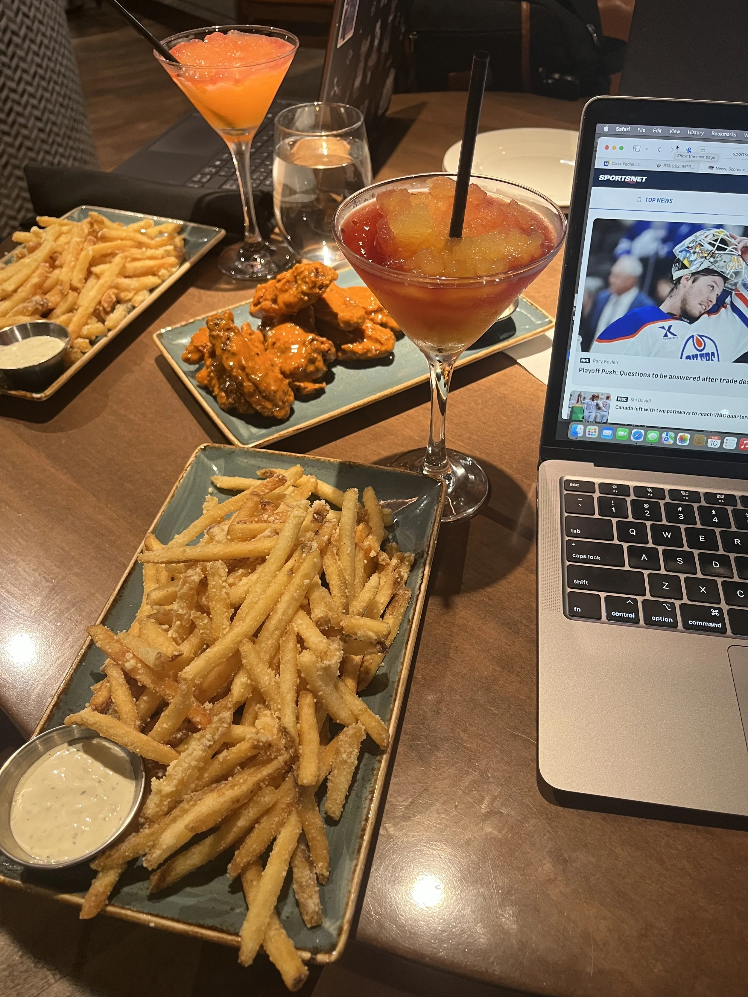 A table with various foods and drinks, including French fries with dipping sauce, chicken wings, and cocktails, alongside a laptop displaying a sports news website.