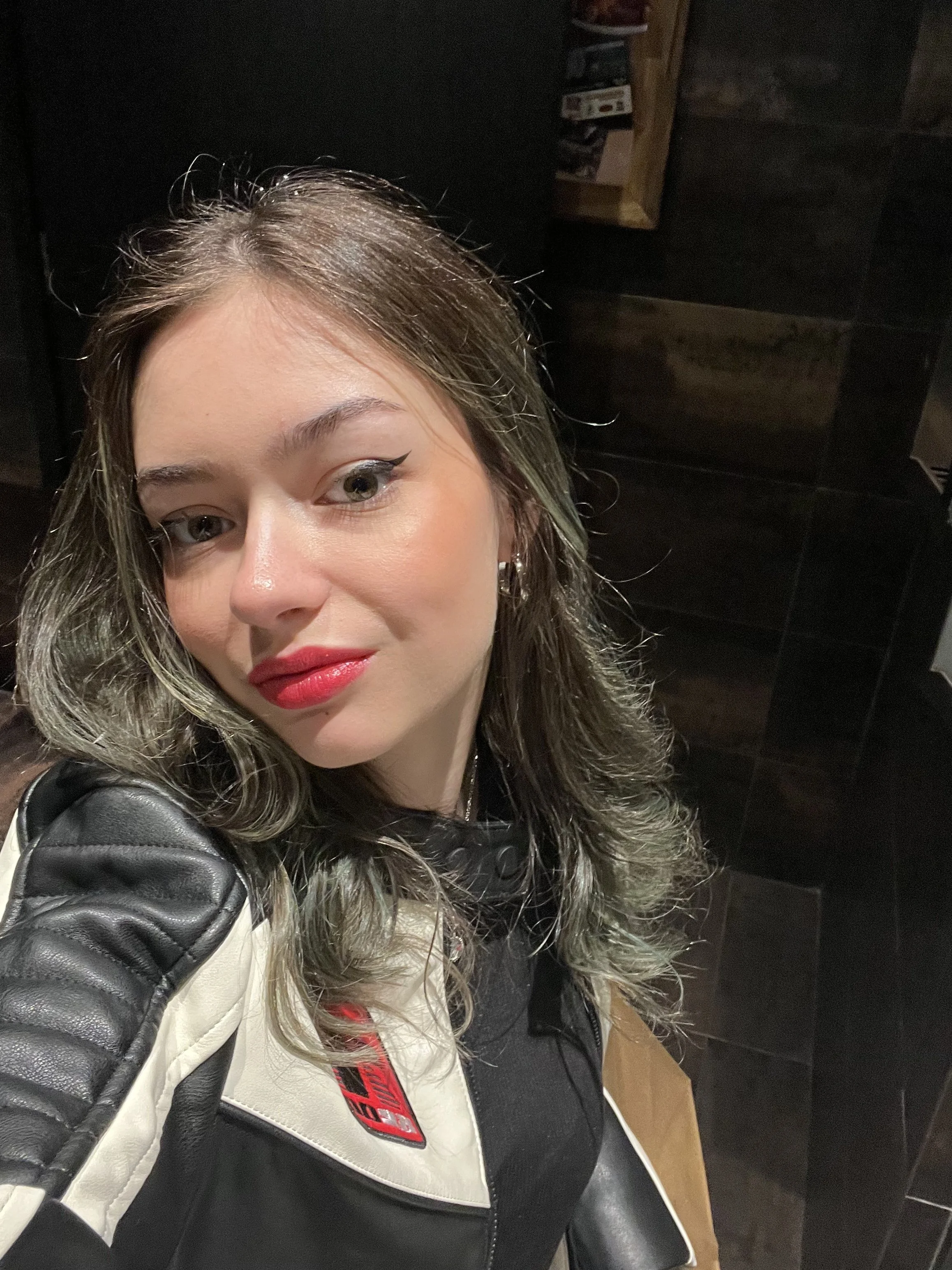 A woman with shoulder-length wavy hair, wearing red lipstick, a leather jacket with a racing stripe, and earrings, taking a selfie in a dimly lit room with dark wood walls.