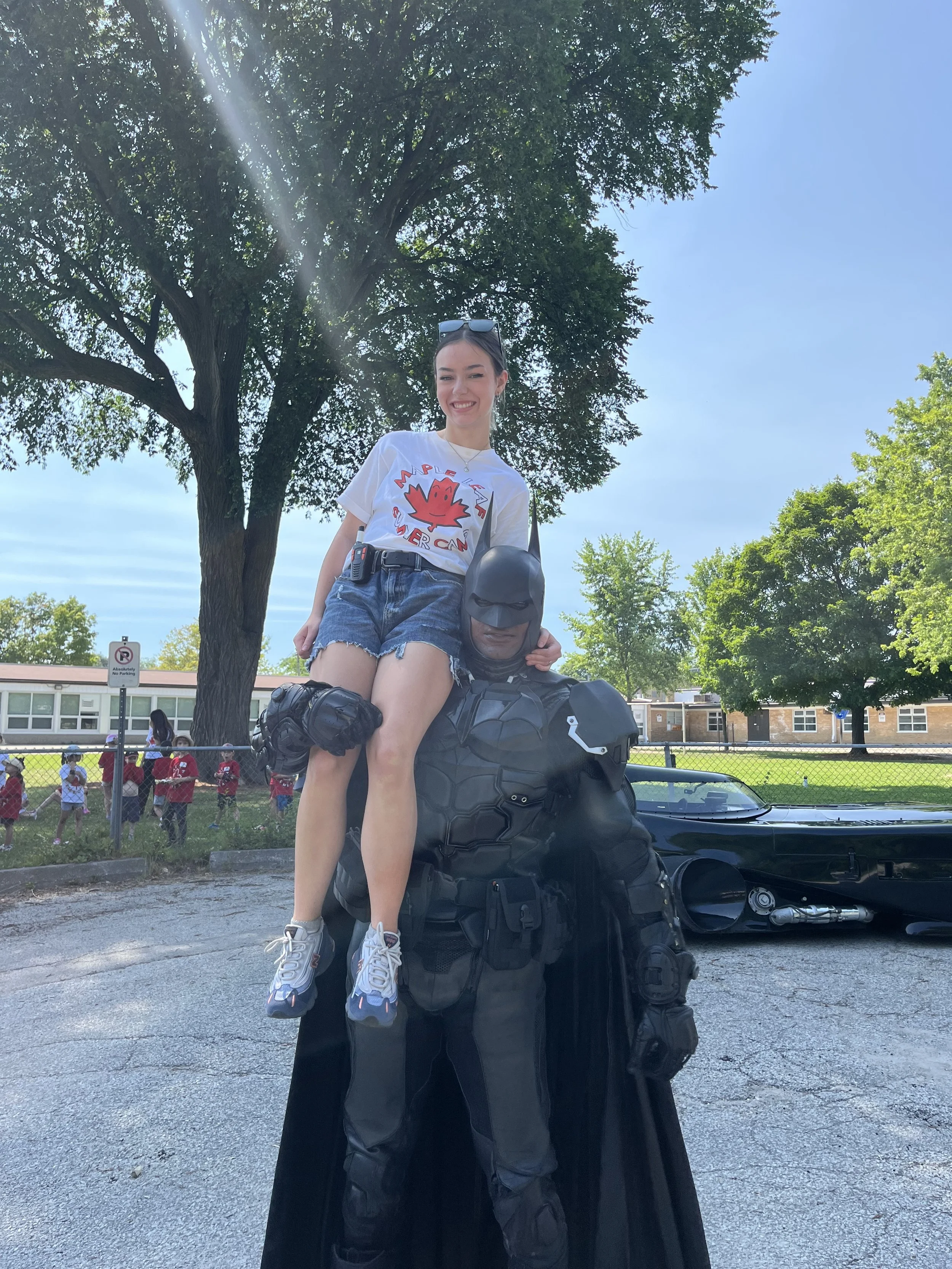 A young woman smiling and wearing sunglasses is sitting on the shoulders of a person dressed as Batman, outdoors near trees and a school building.