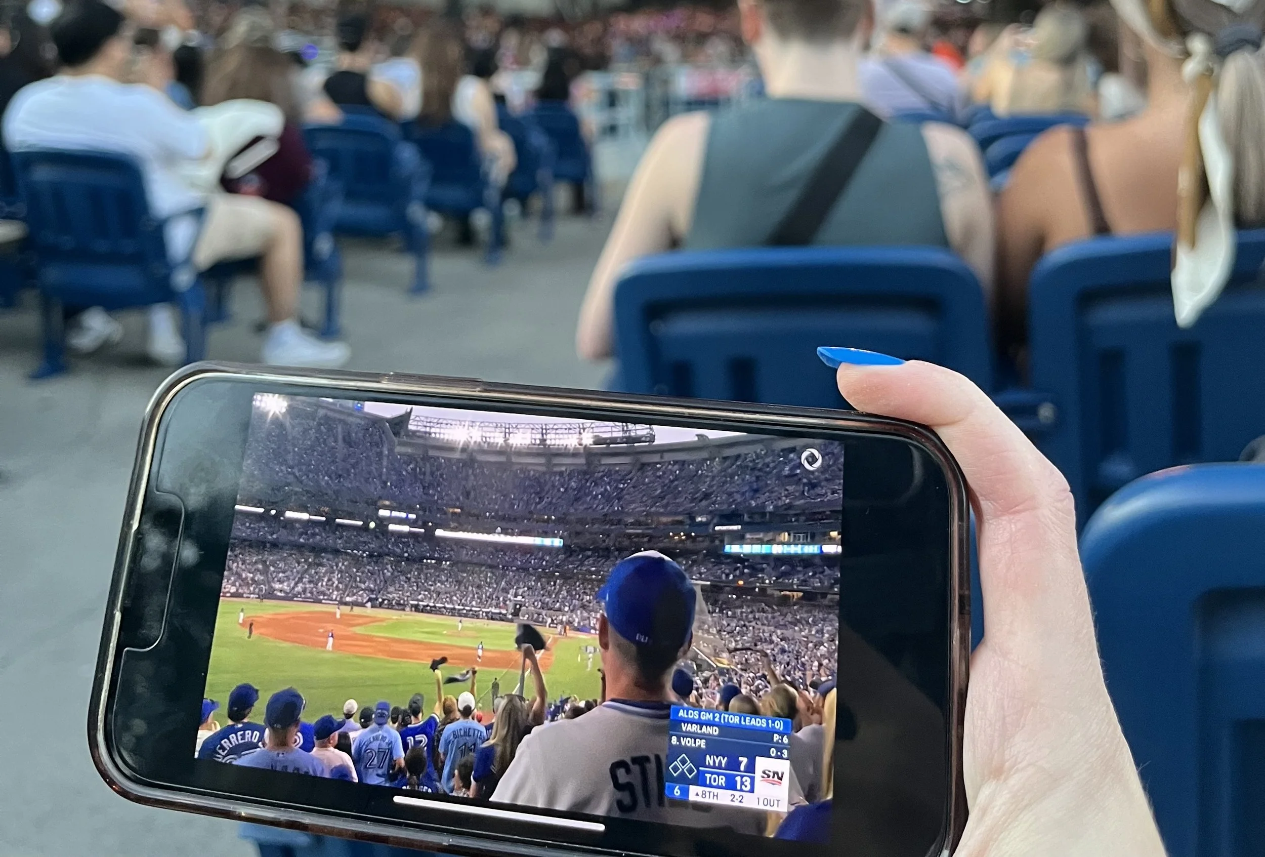 Person watching a baseball game on their phone at a stadium with other fans seated around.