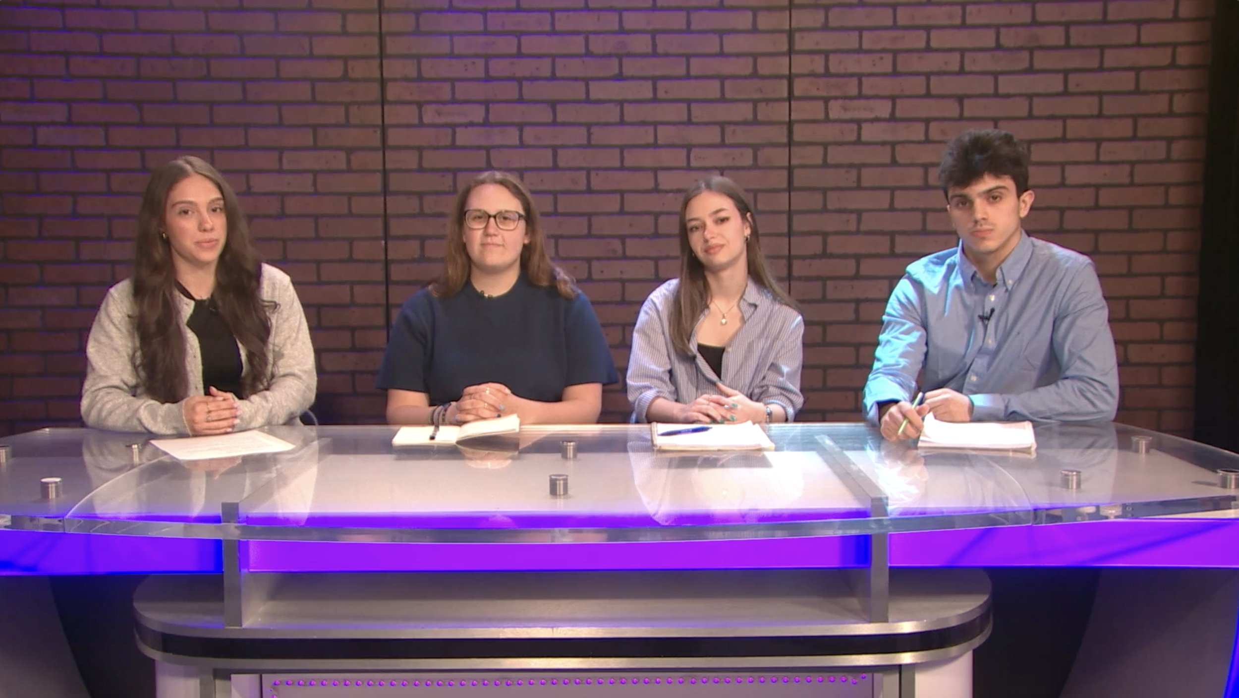 Four young adults sitting at a desk with notebooks and pens, against a brick wall background, appearing to be in a studio or panel setting.