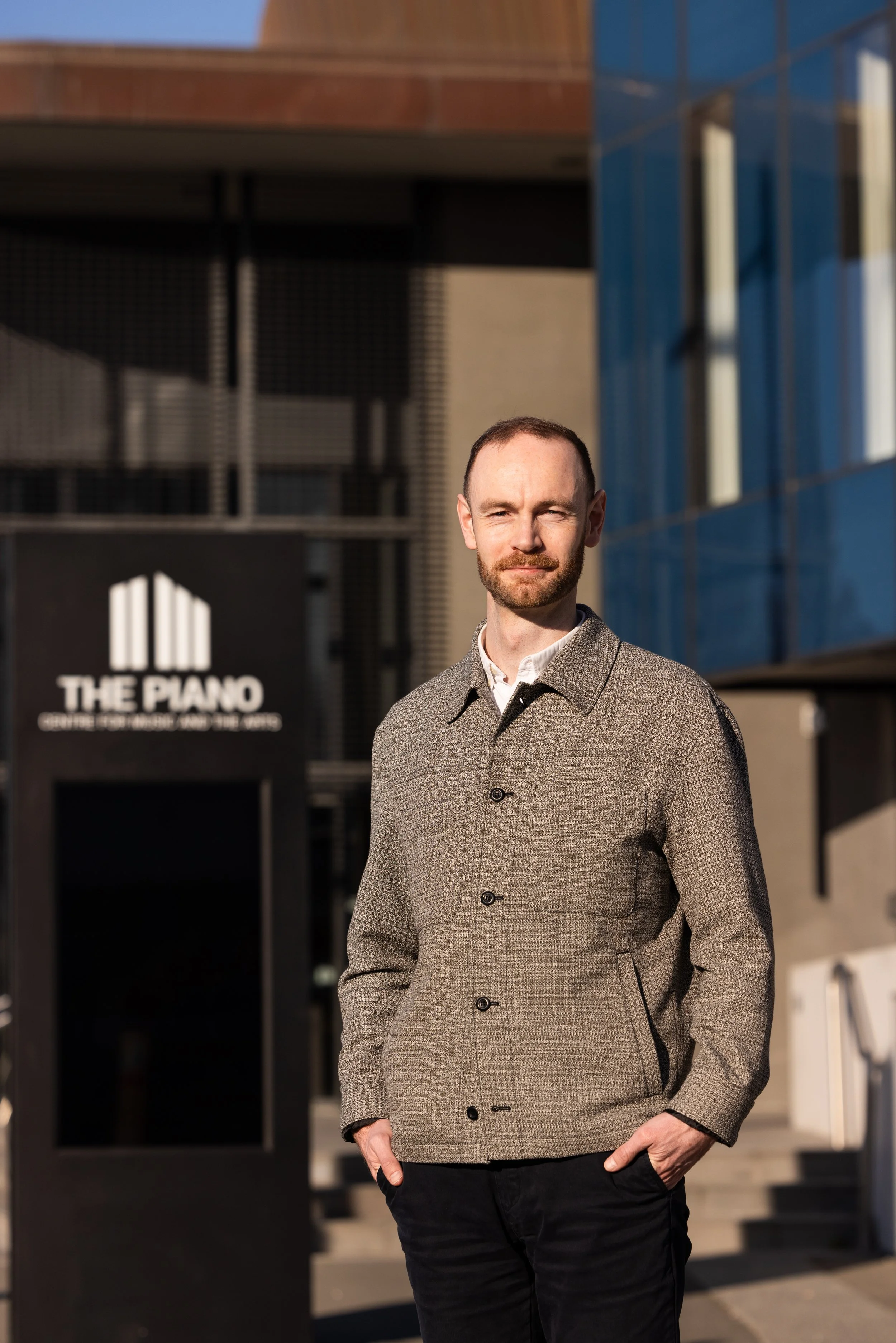 A man with a beard and short hair wearing a gray checkered blazer over a white shirt, standing outdoors in front of modern buildings with a sign that reads 'The Piano Center for Music and the Arts'.