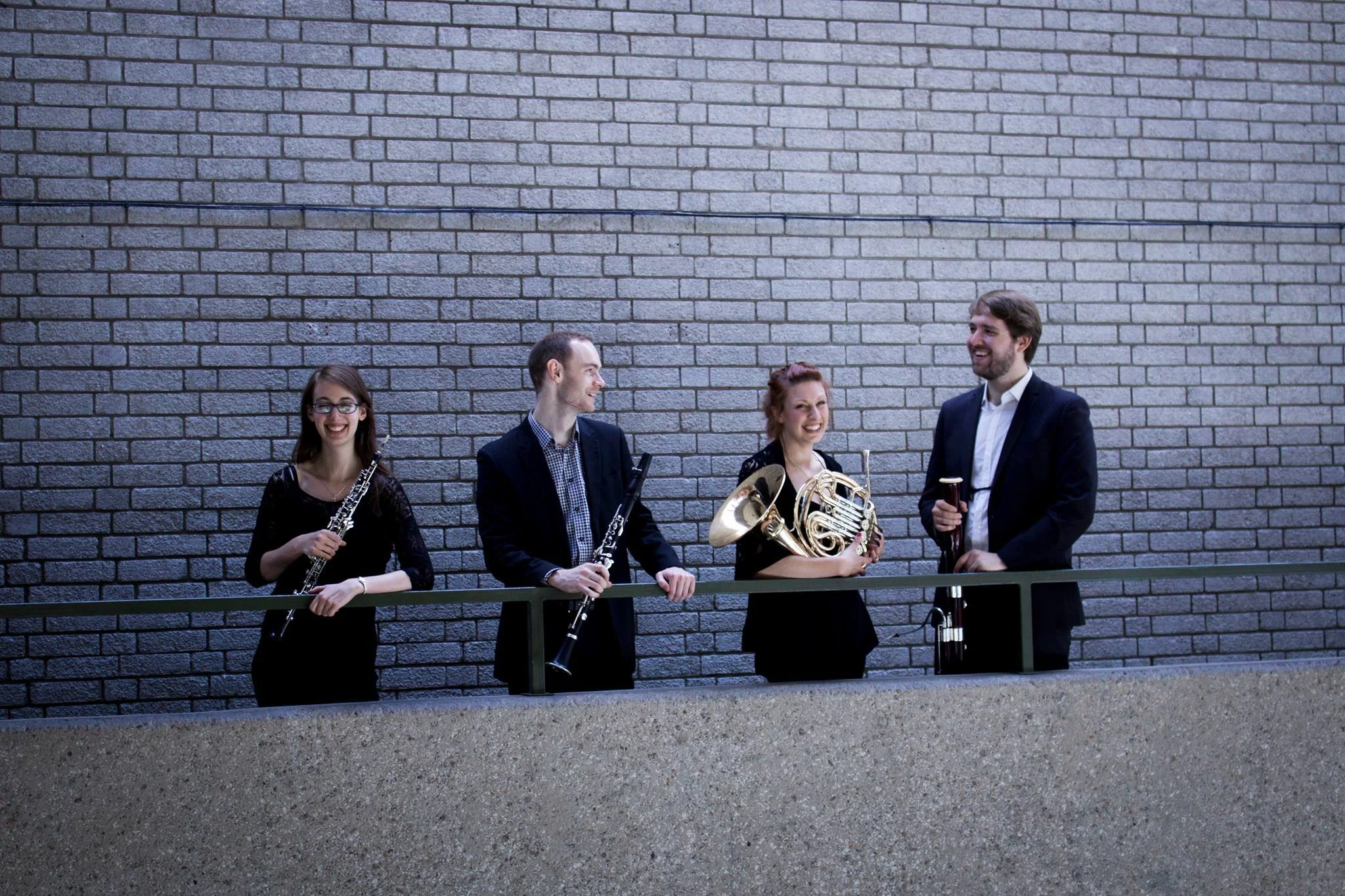 Four musicians in formal attire standing against a brick wall, each holding a musical instrument and smiling, engaged in a conversation.