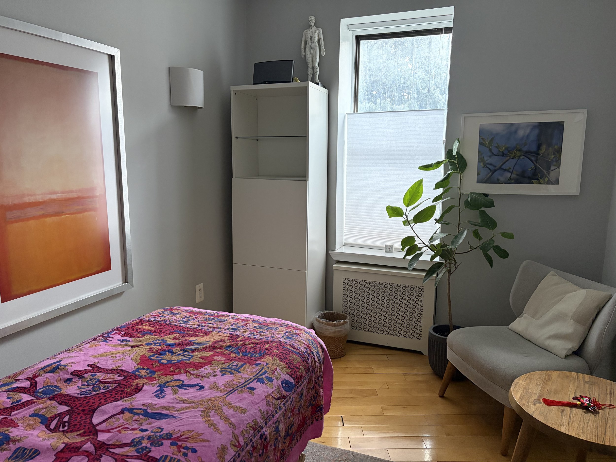 Treatment room  with a bed covered in a colorful pink, purple, and blue patterned quilt, a gray armchair with pillows, a white cabinet with a statue of a human figure on top, a window with natural light, artwork on the walls, and a plant.