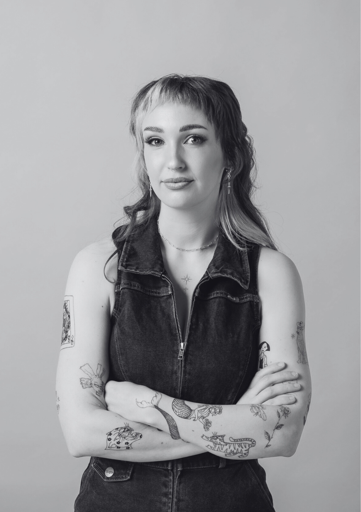 Black and white portrait of a woman with short hair, tattoos on her arms, wearing a sleeveless denim vest and crossing her arms.
