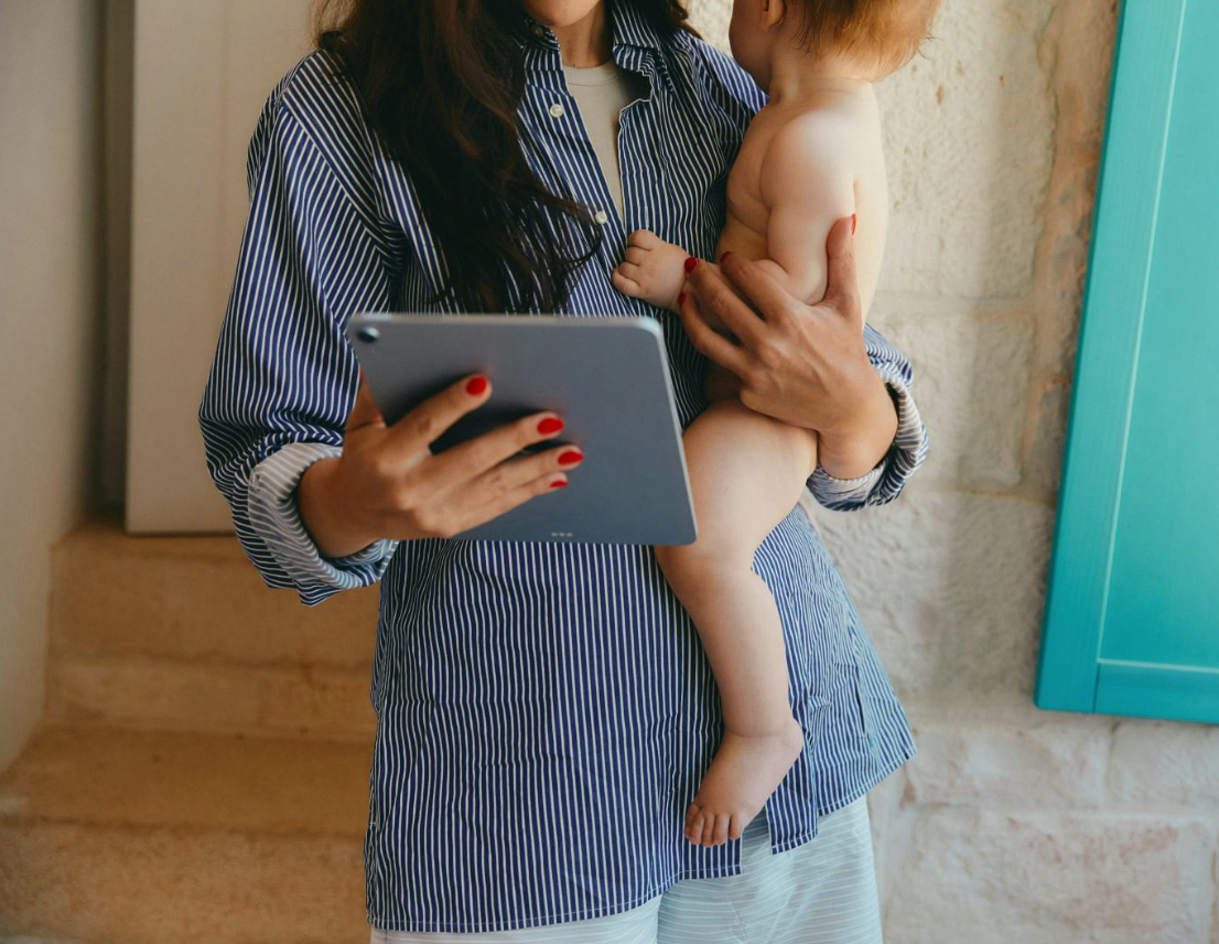 A woman holding a naked toddler while looking at a tablet.