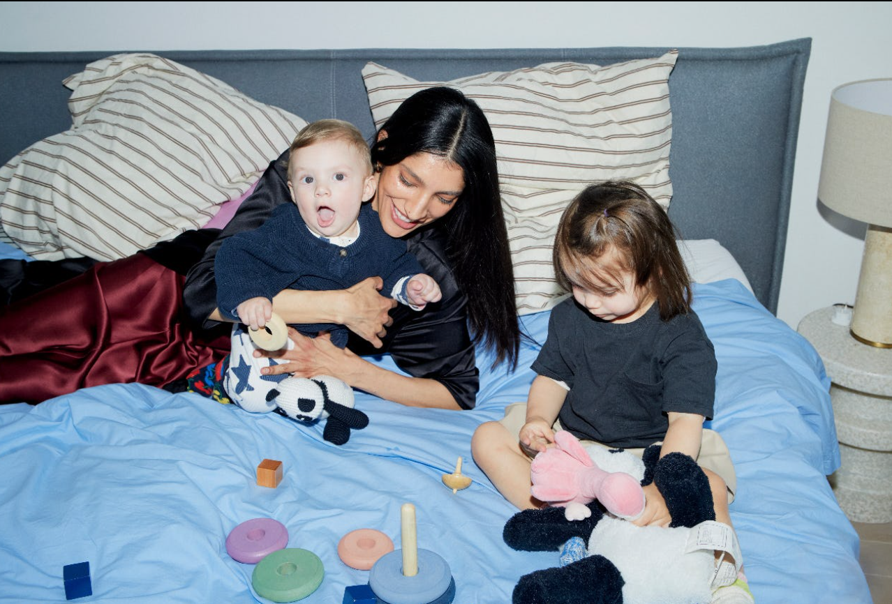 A woman with two children playing on a blue bed with toys, pillows, and a lamp in the background.