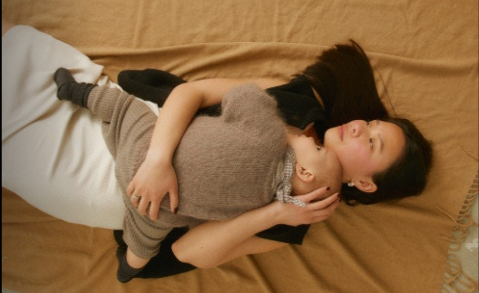 Woman lying on a bed with a young child resting on her chest, both looking content and relaxed.