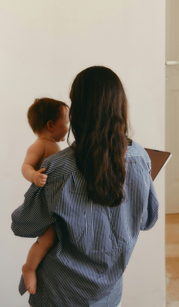 A woman with long brown hair holding a young child with short brown hair, standing indoors near a white wall and looking at a tablet device.