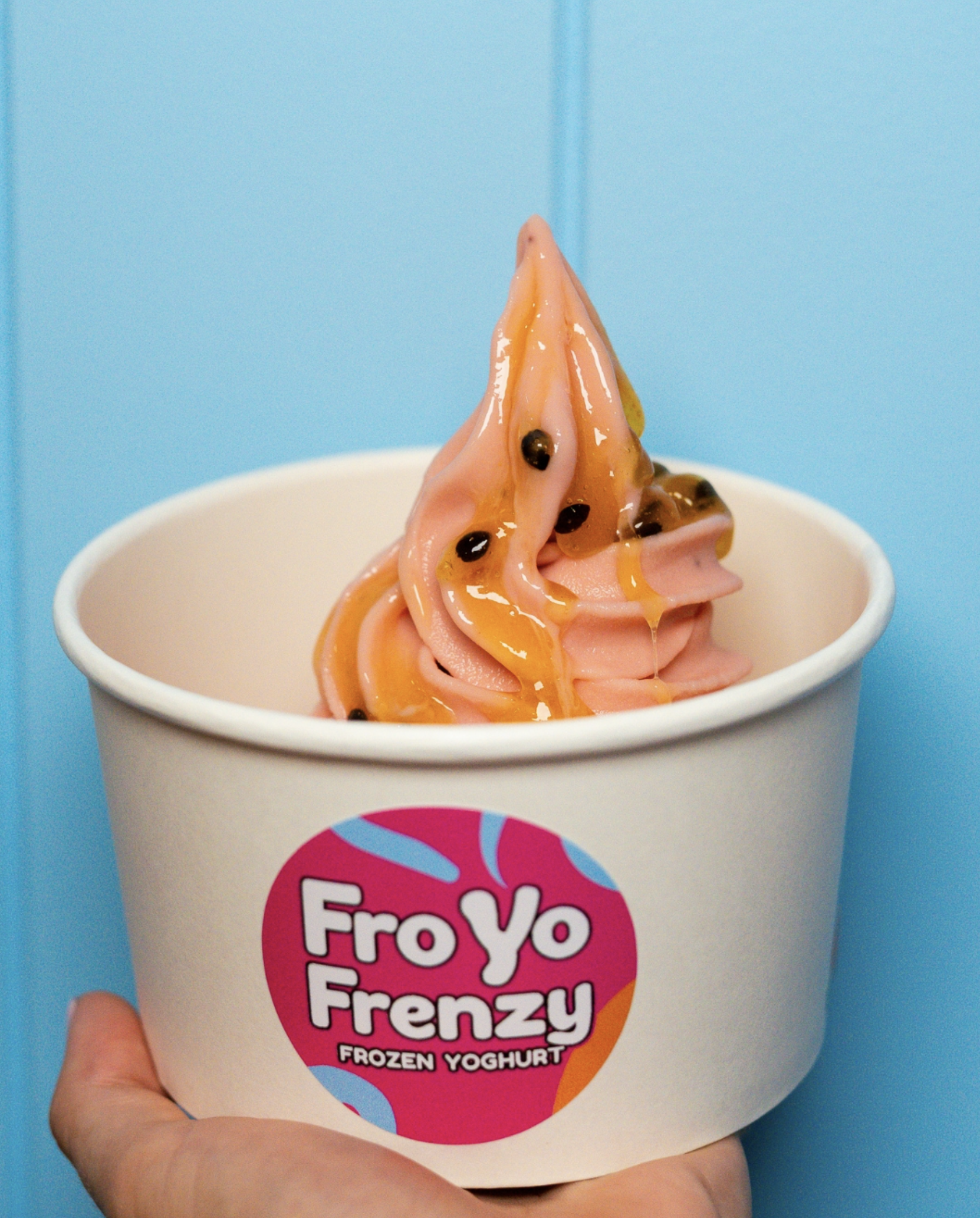 A cup of pink frozen yogurt topped with caramel syrup and passion fruit seeds, held by a hand against a light blue background.