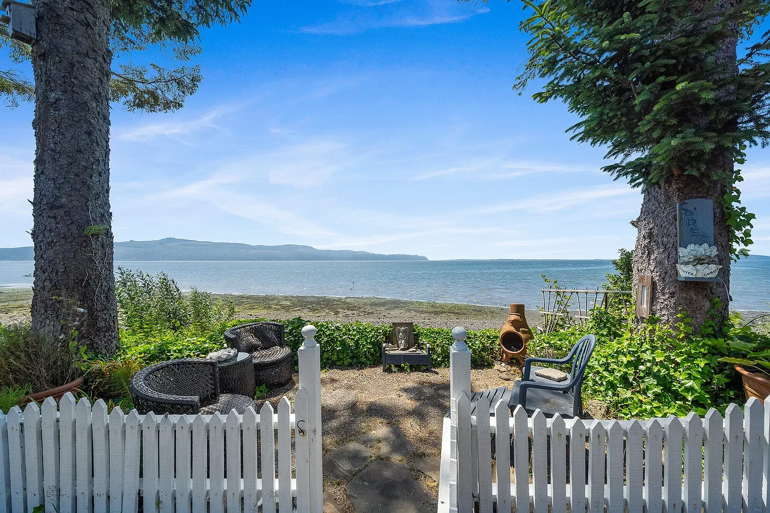 View of a beach from a backyard with a white picket fence, outdoor chairs, and a fire pit, overlooking a calm sea with distant hills under a blue sky.