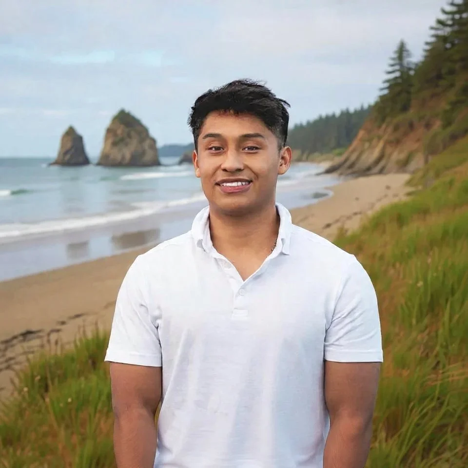 A young man smiling at the camera, standing on a beach with sand, ocean waves, rocky formations in the water, and cliffs with trees in the background.