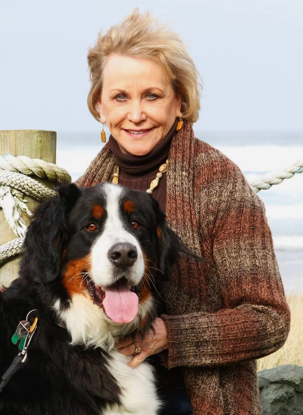 A smiling woman with short blonde hair wearing a brown sweater and scarf, standing outdoors with a Bernese mountain dog in front of her near a wooden post with a thick rope, against a cloudy sky.