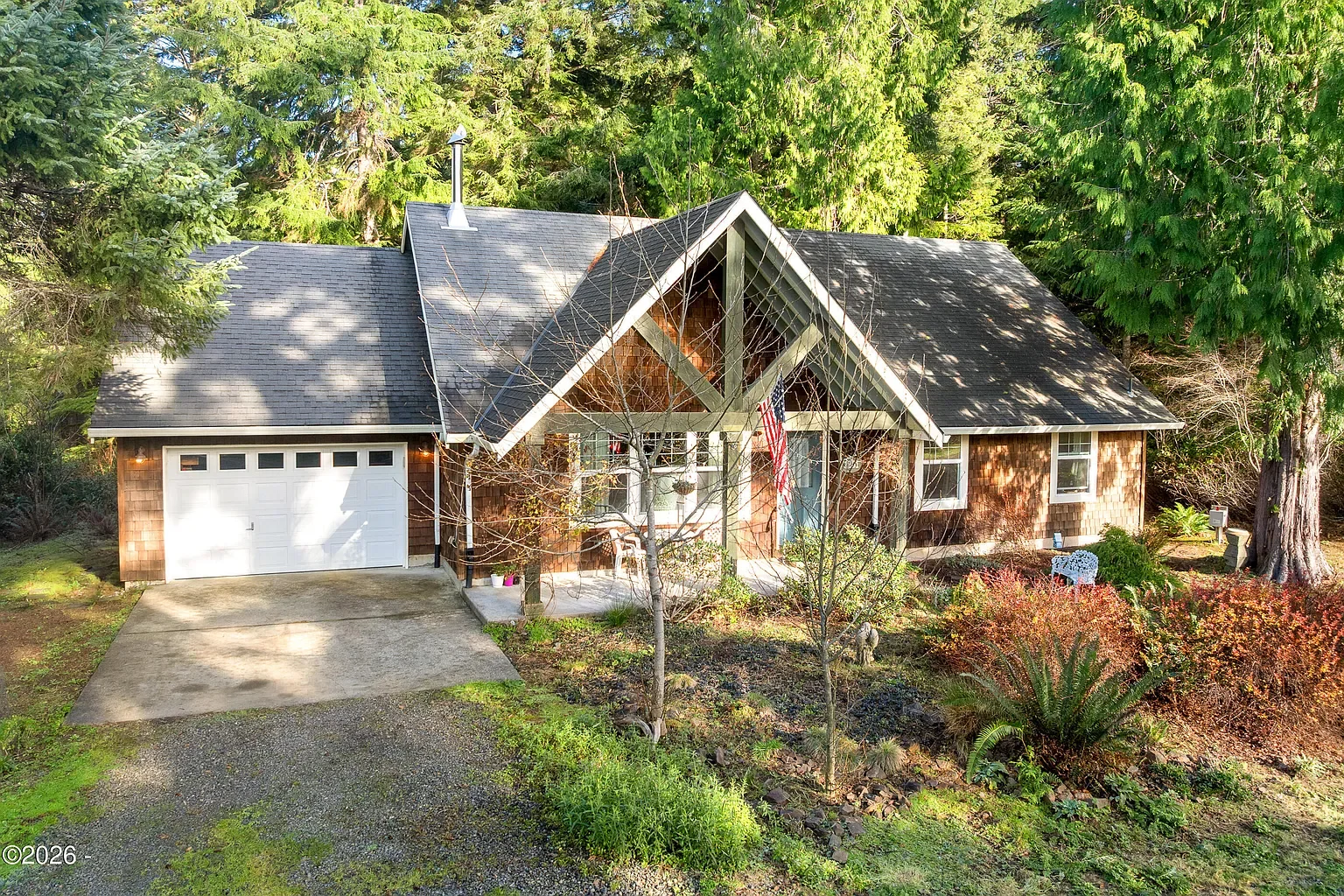 A single-story house with a triangular front porch, dark shingle roof, and attached garage, surrounded by trees and a garden in a wooded setting.
