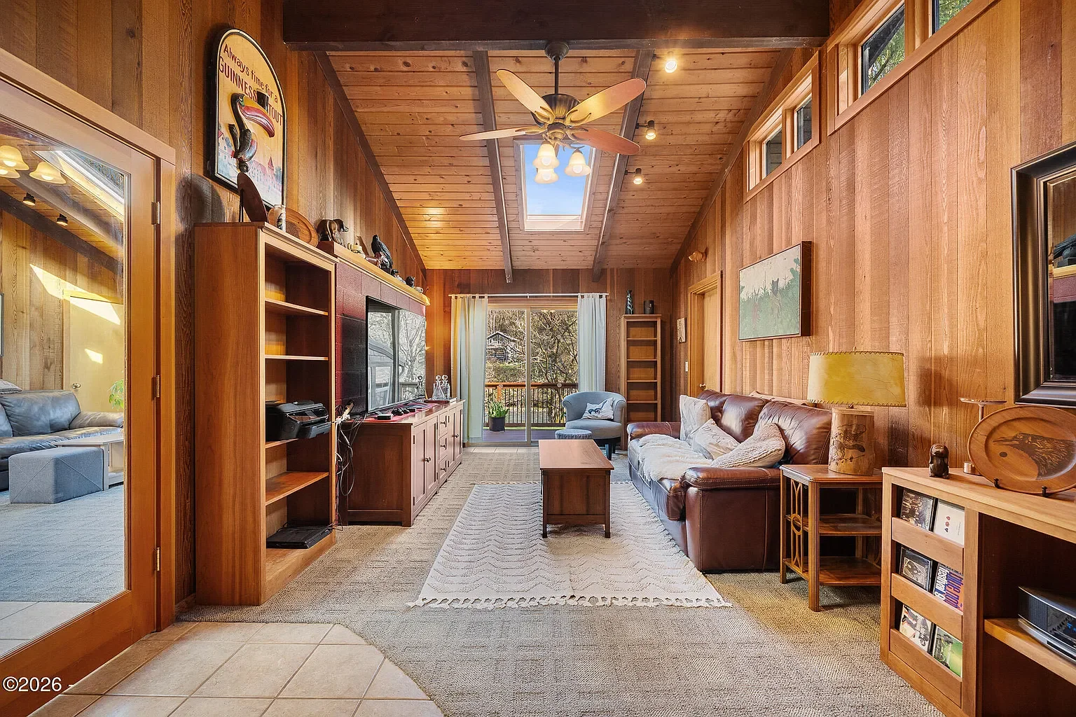 Living room with wooden walls and ceiling, ceiling fan, skylight, window with curtains, brown leather sofa, armchair, wooden furniture, TV, and a balcony door leading outside.