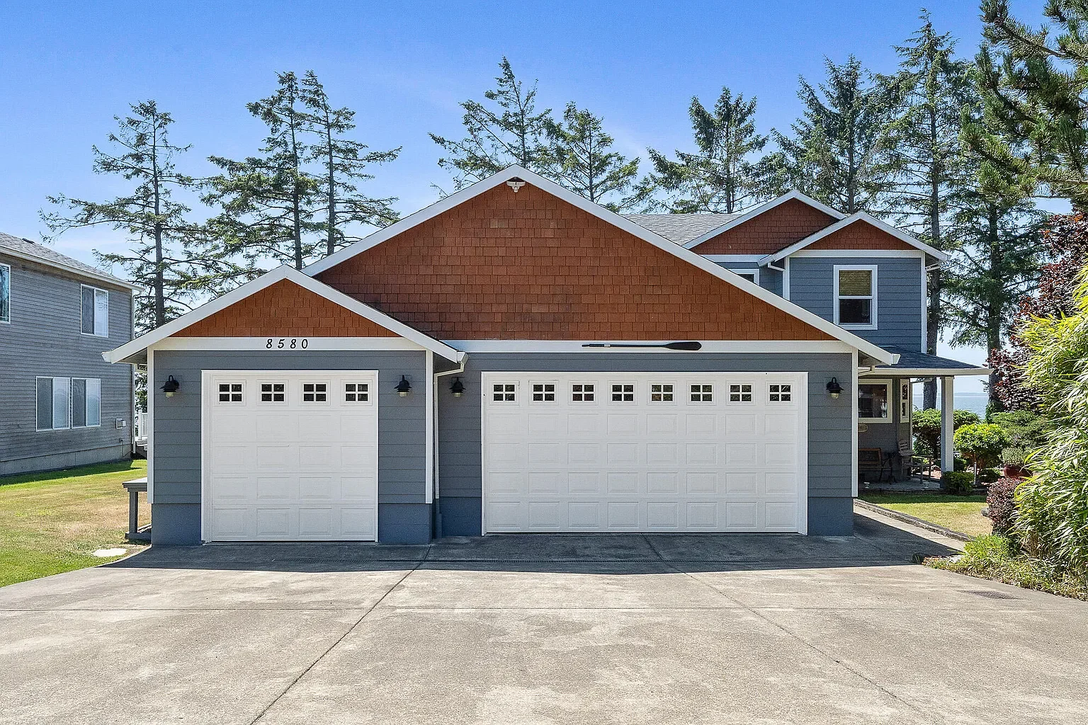 Front view of a two-story house with a double garage, light blue siding, and brown shingle accents, surrounded by trees and a well-maintained yard.