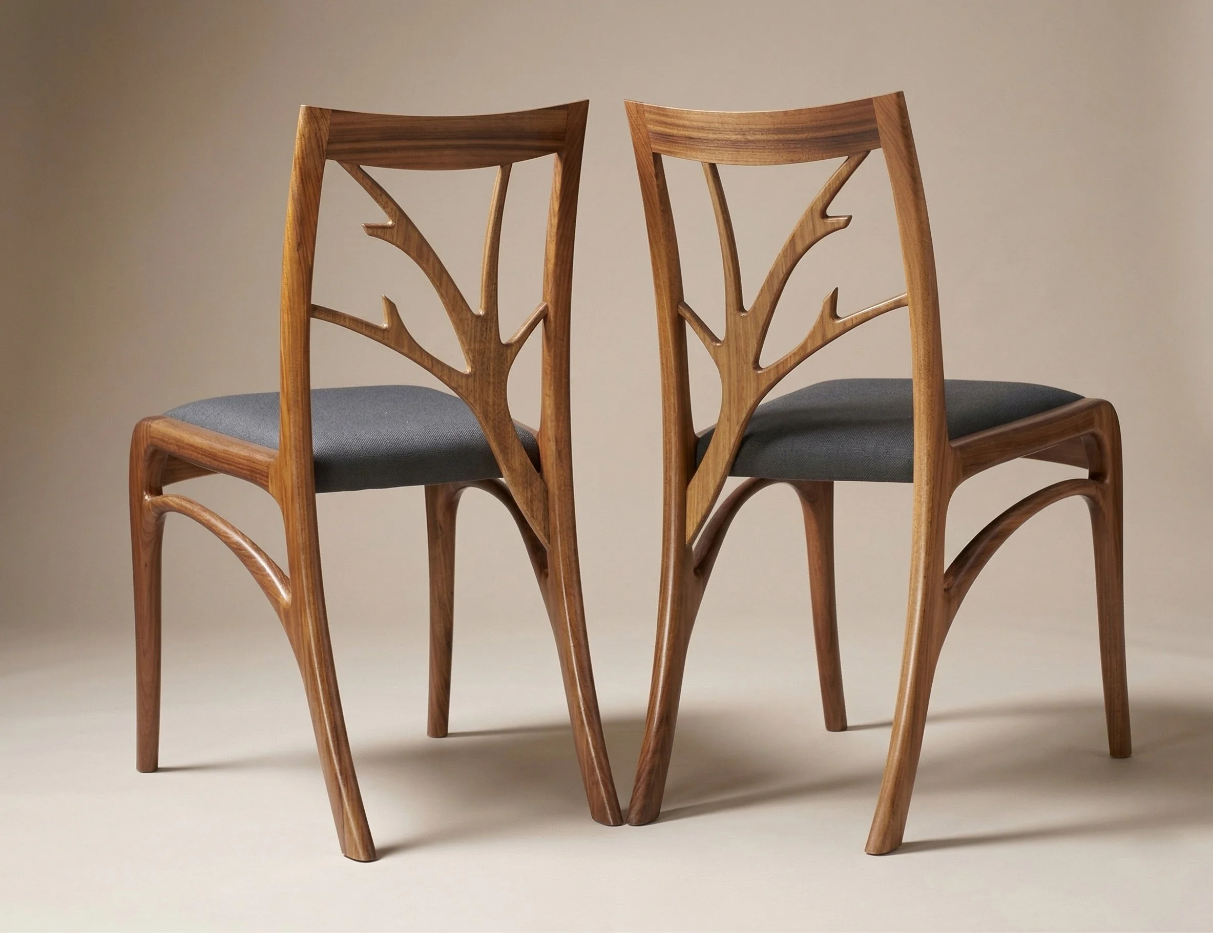 Two wooden custom made dining chairs with hand-shaped tree branch-shaped backrests and dark gray upholstery cushioned seats are placed side by side against a plain wall. Handmade in Brisbane Australia from Tasmania Blackwood by Will Marx