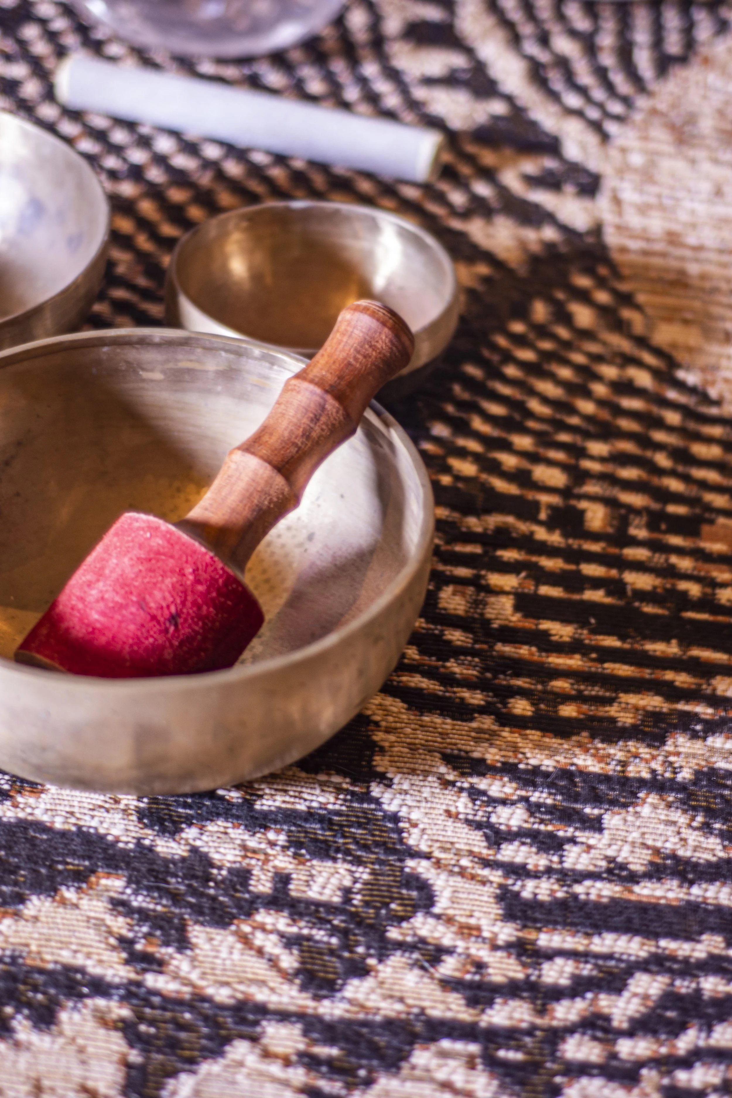 A singing bowl with a wooden striker inside, on a patterned woven surface with other small bowls and a white stick nearby.