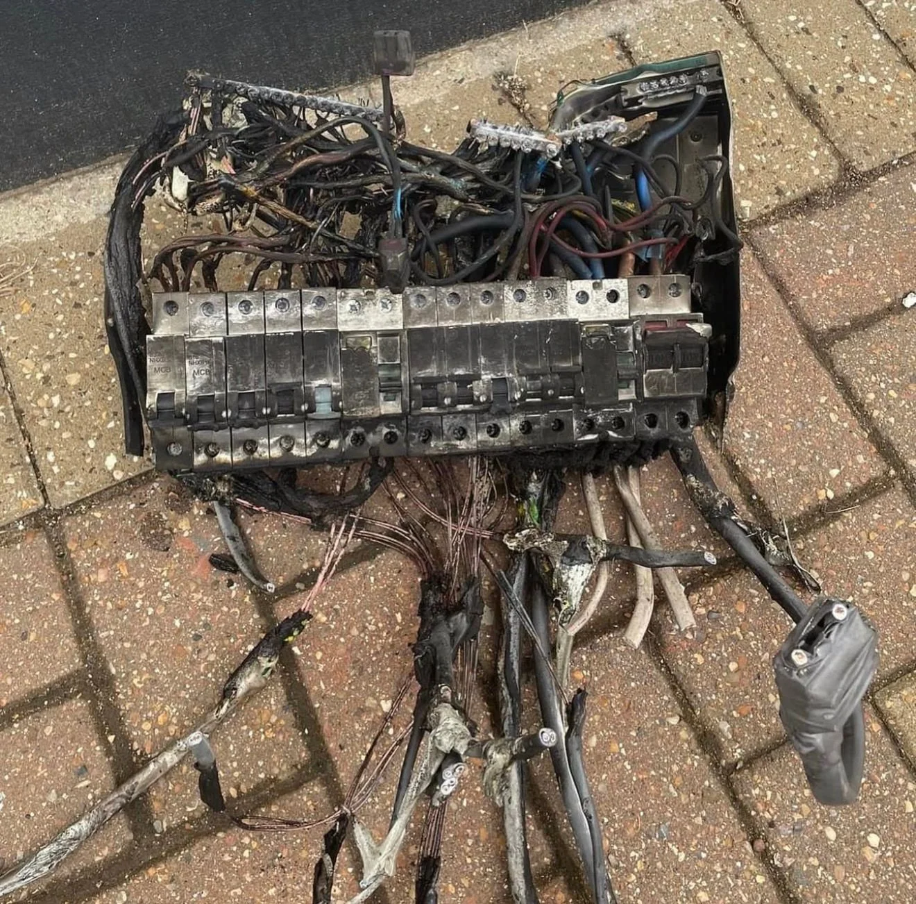 Damaged and burnt electrical wiring and fuse box on brick sidewalk.