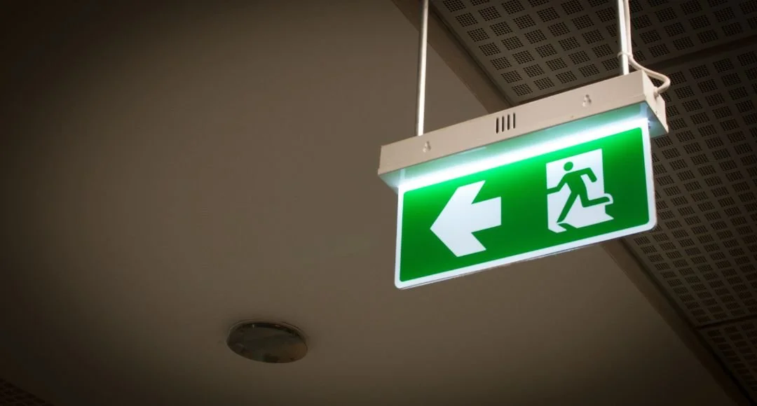 Green emergency exit sign with a running person and an arrow pointing left, mounted on the ceiling.