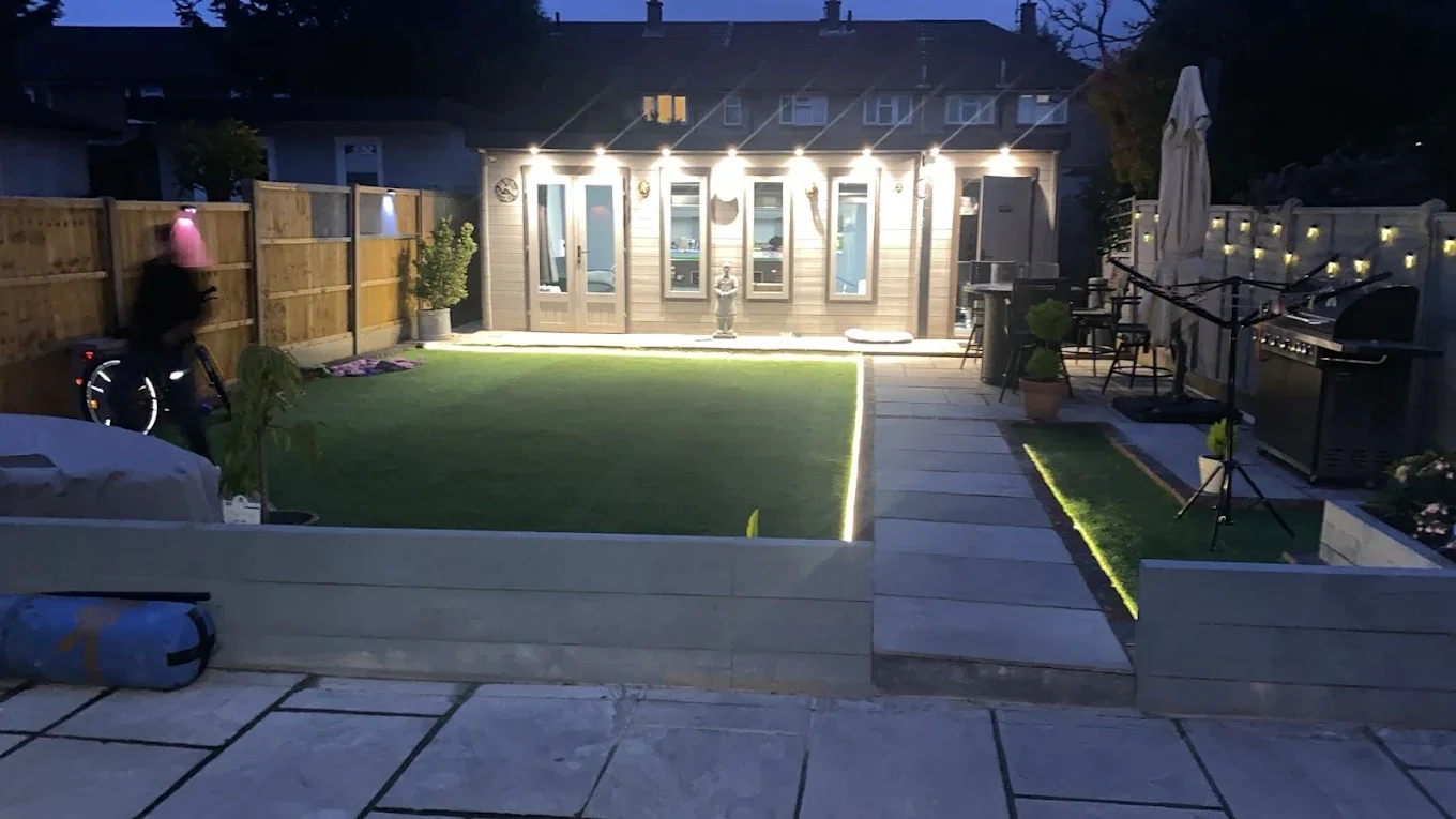 A backyard at dusk with a well-lit house, a small lawn, a paved patio area with outdoor furniture, string lights along the fence, and a person with a pink hoodie walking a bicycle.