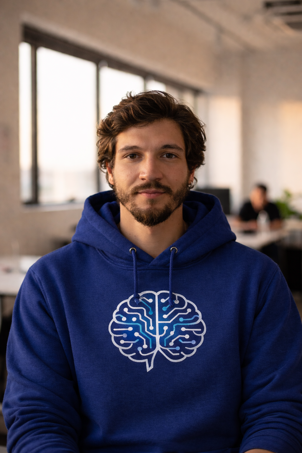 A young man with brown hair, facial hair, and light skin, wearing a blue hoodie with a white and blue brain illustration on it, sitting indoors with windows behind him and a blurred background.