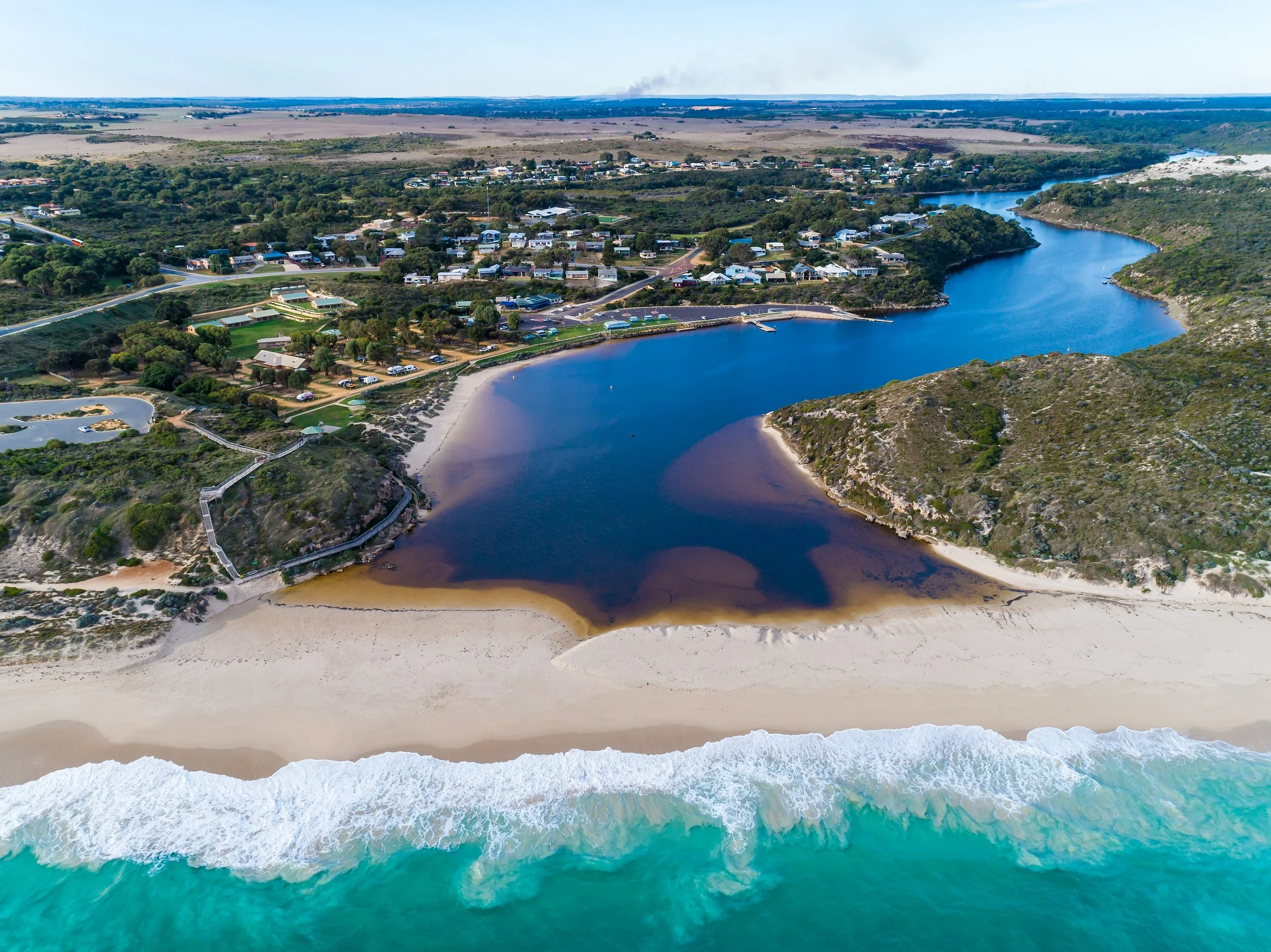 Aerial view of Moore River, Guilderton coastal area with a sandy beach, a dark-colored river, and residential houses along the coastline with Wilbinga hills, trees, and water bodies in the background.