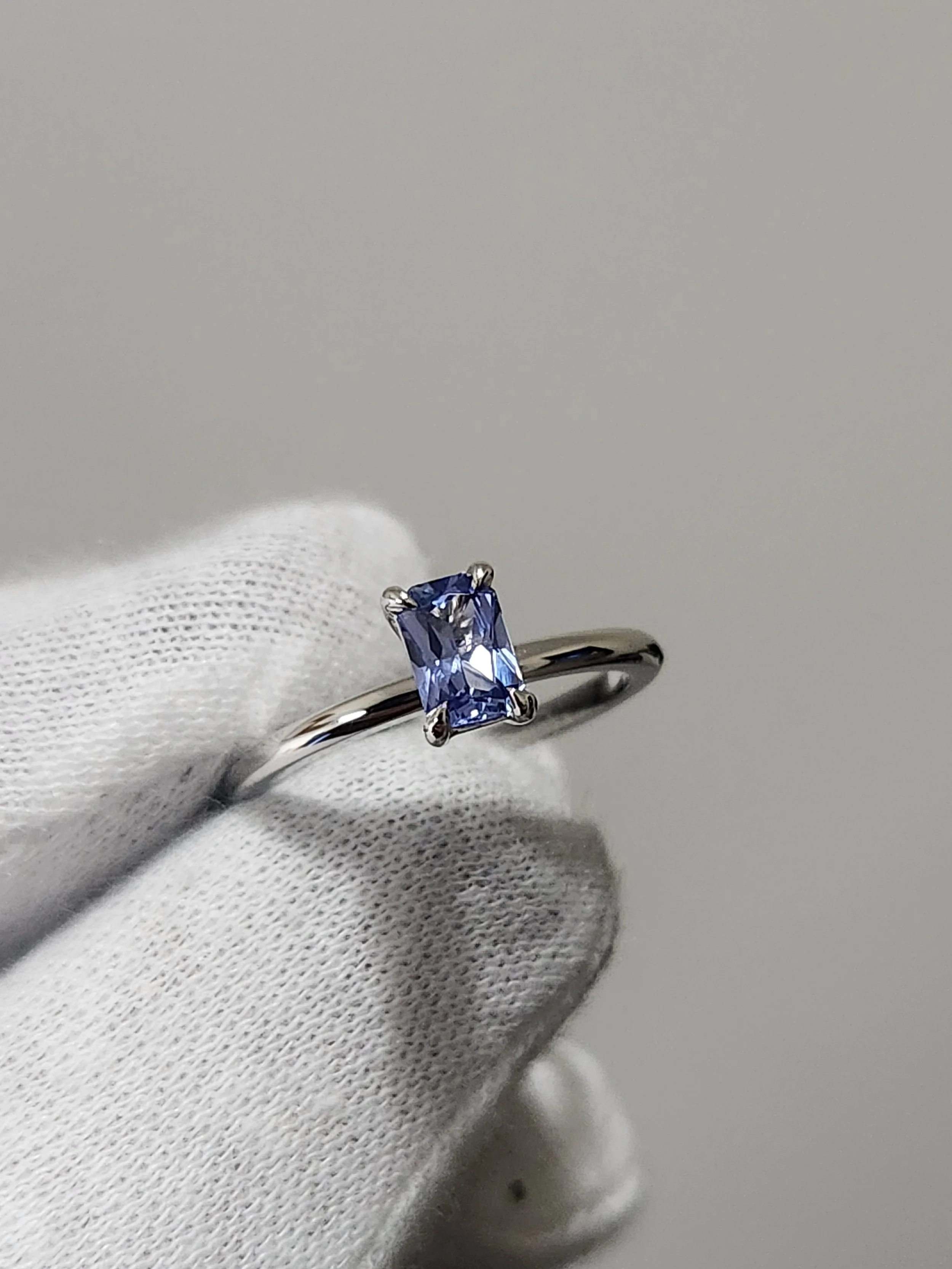 A white metal solitaire style ring with an emerald cut tanzanite gemstone, held by a white gloved hand.