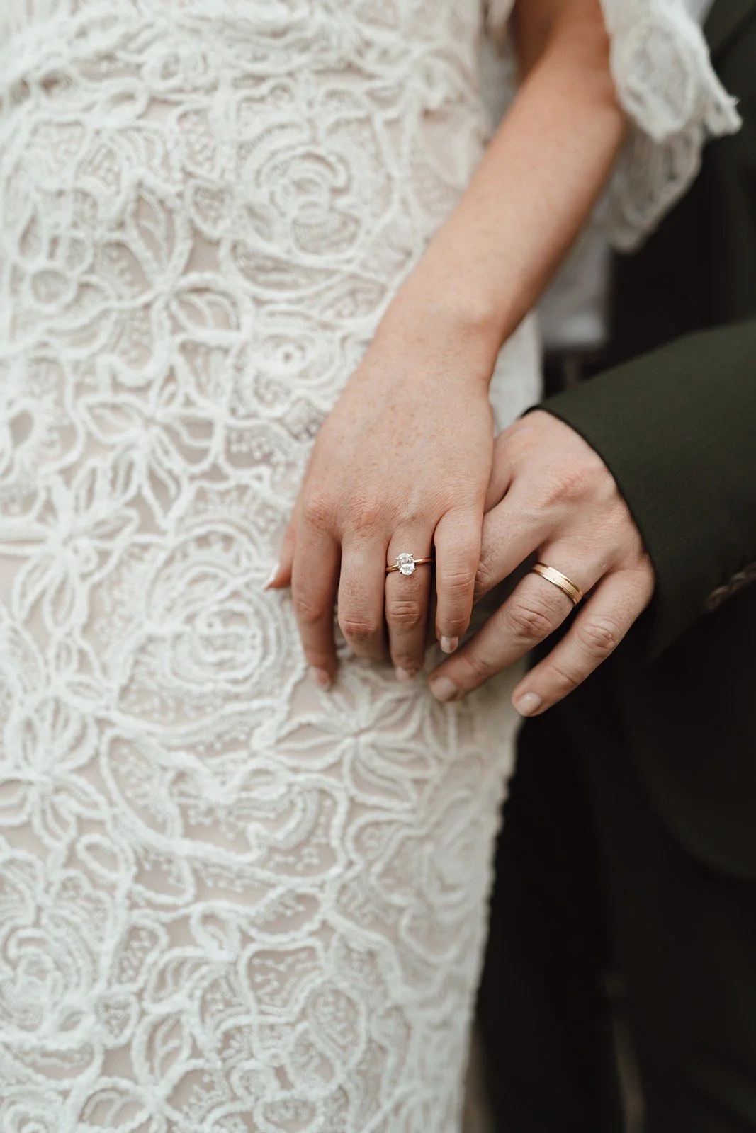 Close-up of a bride and groom holding hands, showing wedding rings, with a white lace dress in the background.
