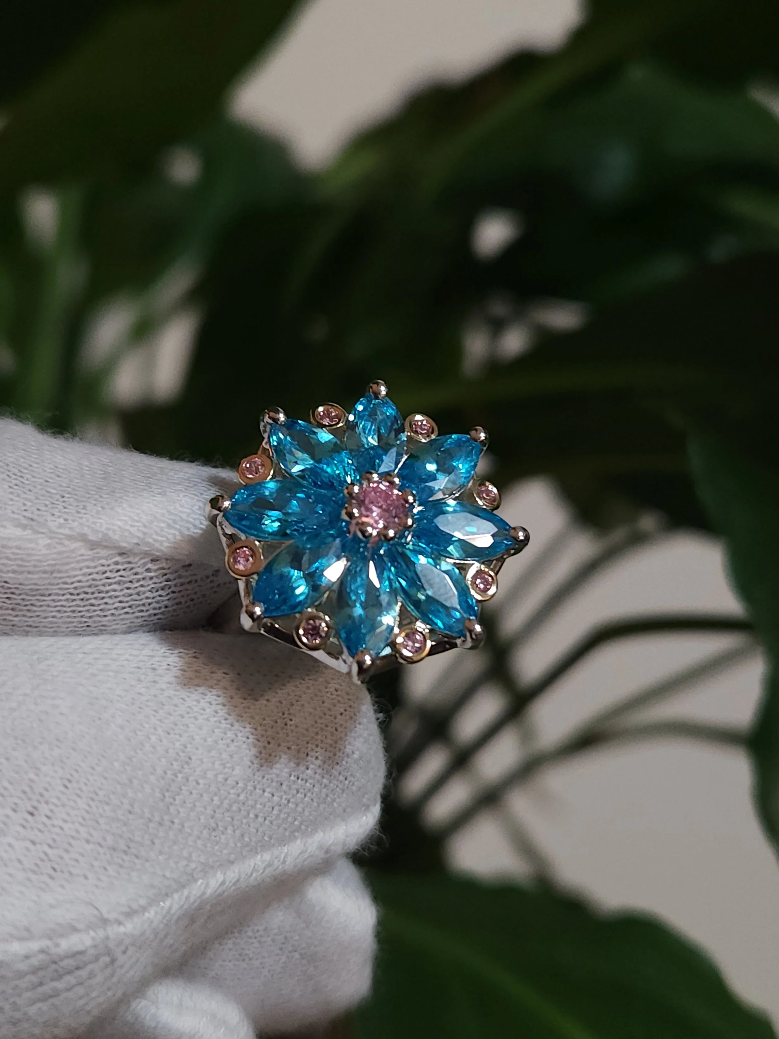 A cluster style white gold ring with a large floral design made of natural blue topaz petals and pink topaz centre, held by a white-gloved hand.