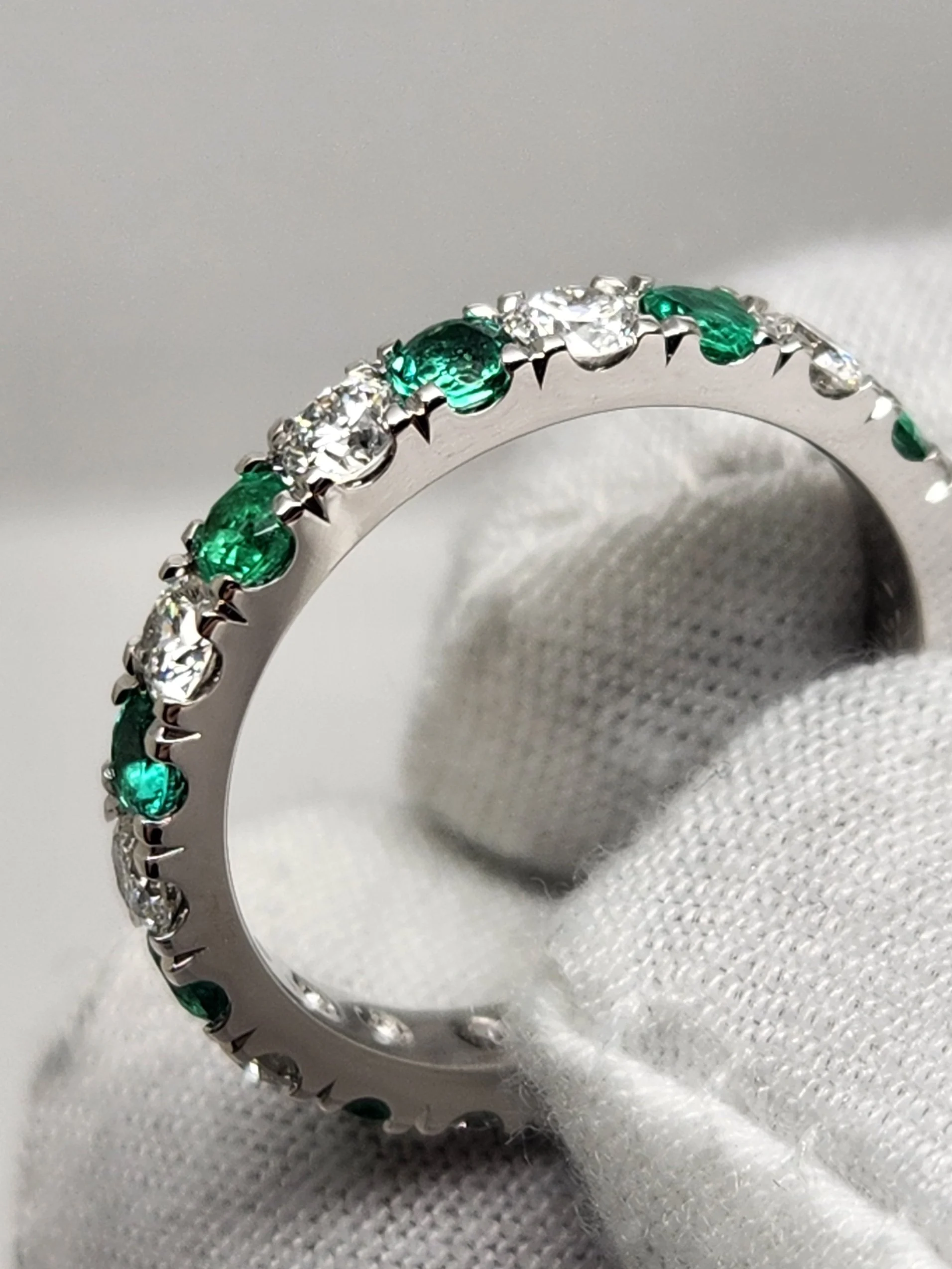 A white metal ring with alternating oval cut natural green emeralds and near-colourless natural diamonds.