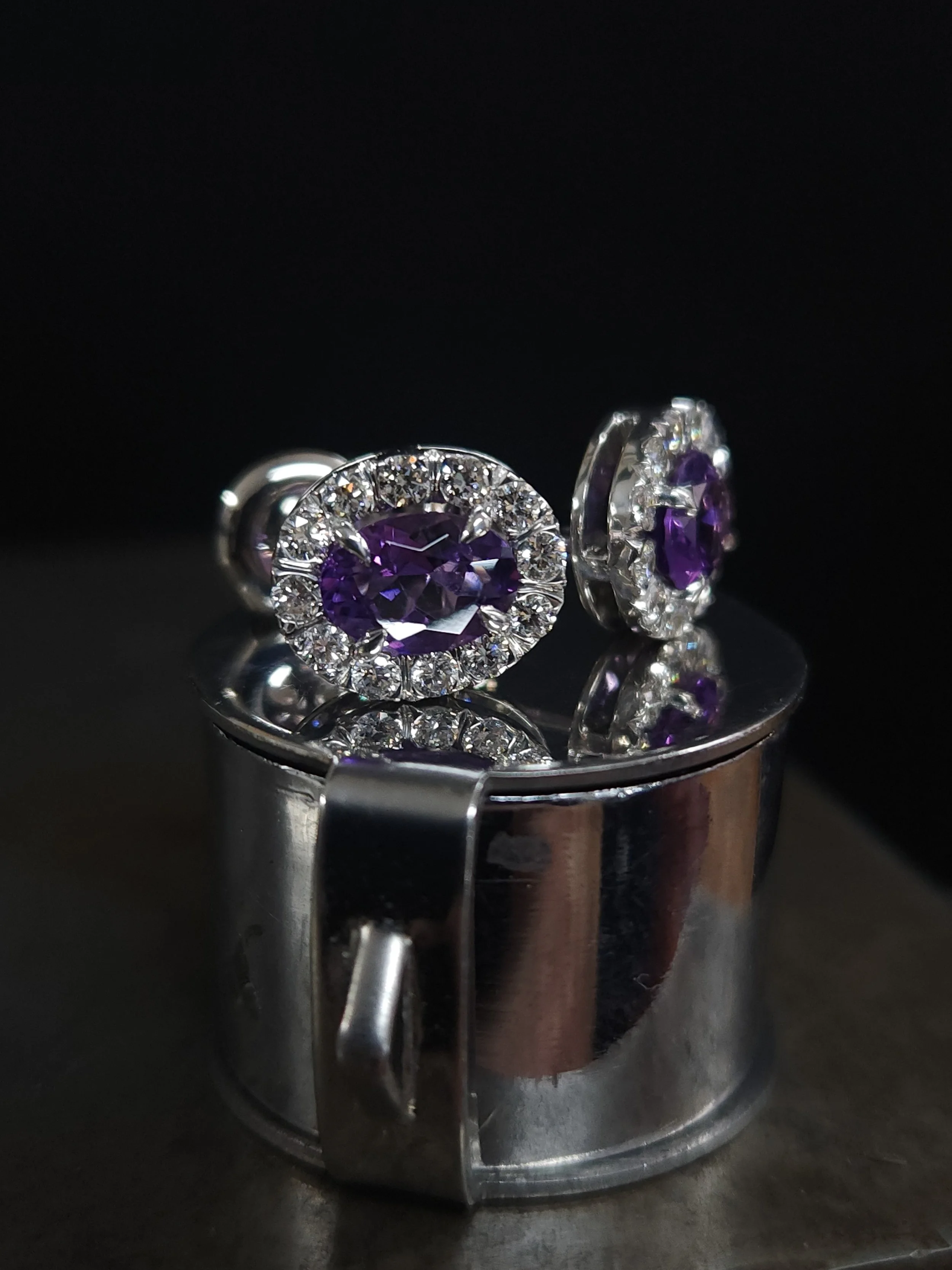 A pair of white gold stud earrings with oval cut purple amethyst gemstones surrounded by round brilliant cut natural near-colourless diamonds in claw settings.