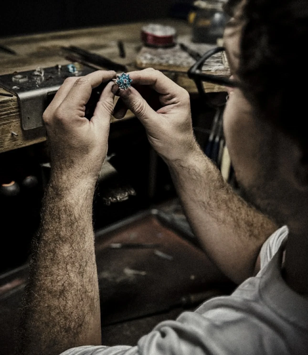 A person, Jordan the Jeweller, with glasses on, working on a ring at a workbench, surrounded by tools and materials.