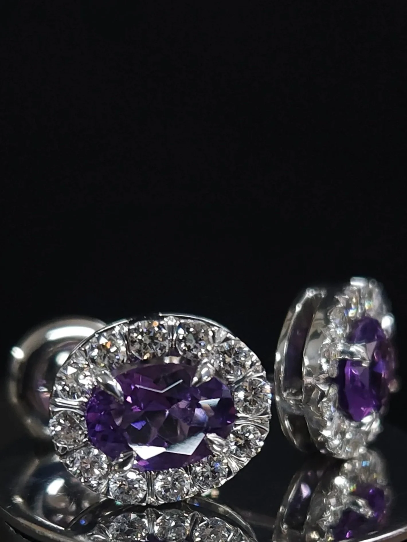 A pair of white gold stud earrings with oval cut purple amethyst gemstones surrounded by round brilliant cut natural near-colourless diamonds in claw settings.