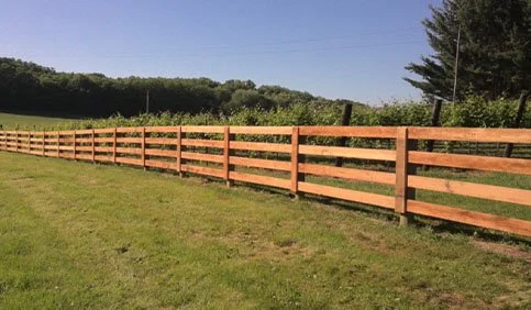 A wooden fence running along a grassy field with trees and a blue sky in the background.