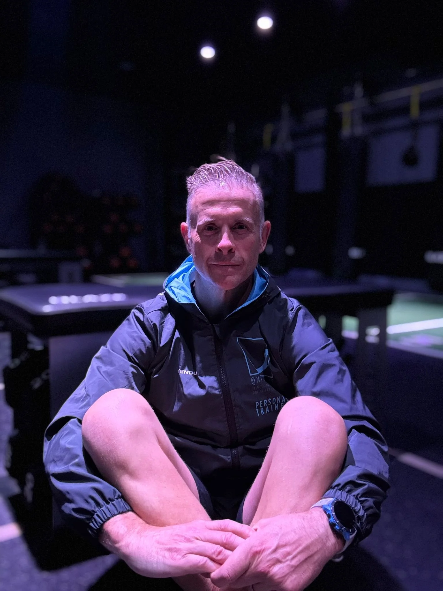 A man sitting with his knees up, wearing a black jacket with 'PERSONAL TRAINER' written on it, in a dimly lit room, possibly a gym or sports facility, with reflections on his face from overhead lights.
