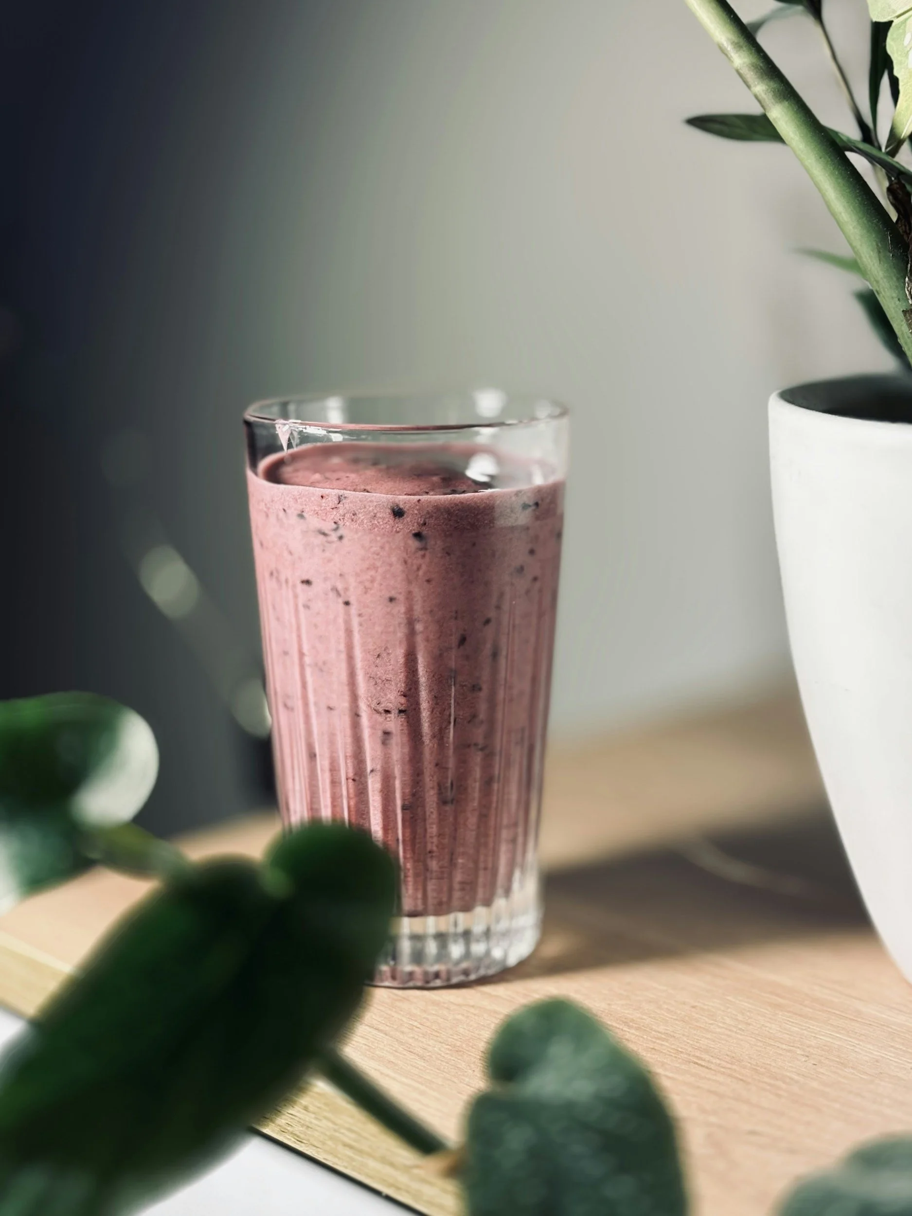 A pink smoothie in a tall, transparent glass with a textured pattern, placed on a wooden surface. In the background, a green leafy plant in a white pot and some out-of-focus green leaves in the foreground.