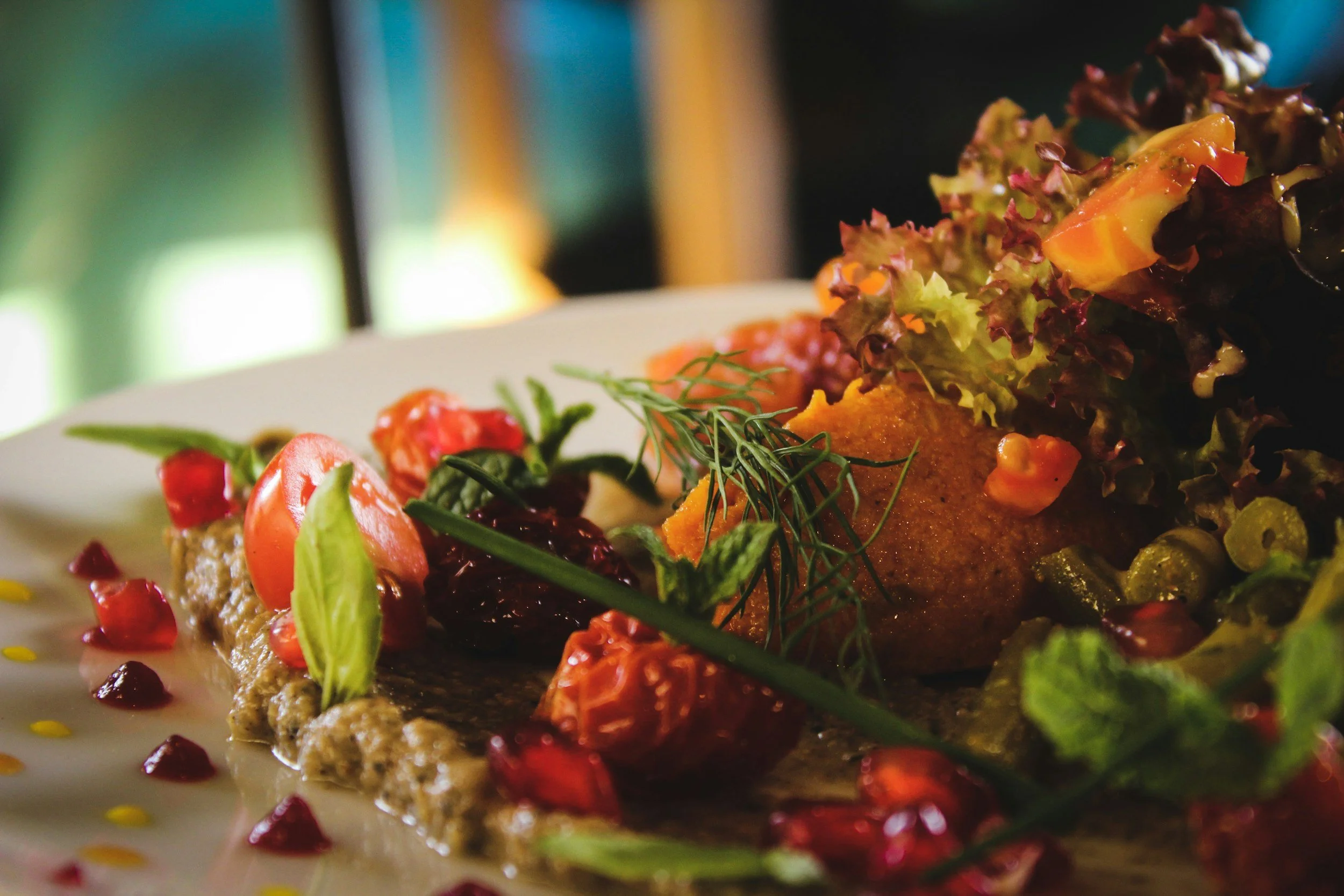 Close-up of a plate with colorful salad and a piece of breaded fried food, garnished with herbs, pomegranate seeds, and dressing, with a blurred background.