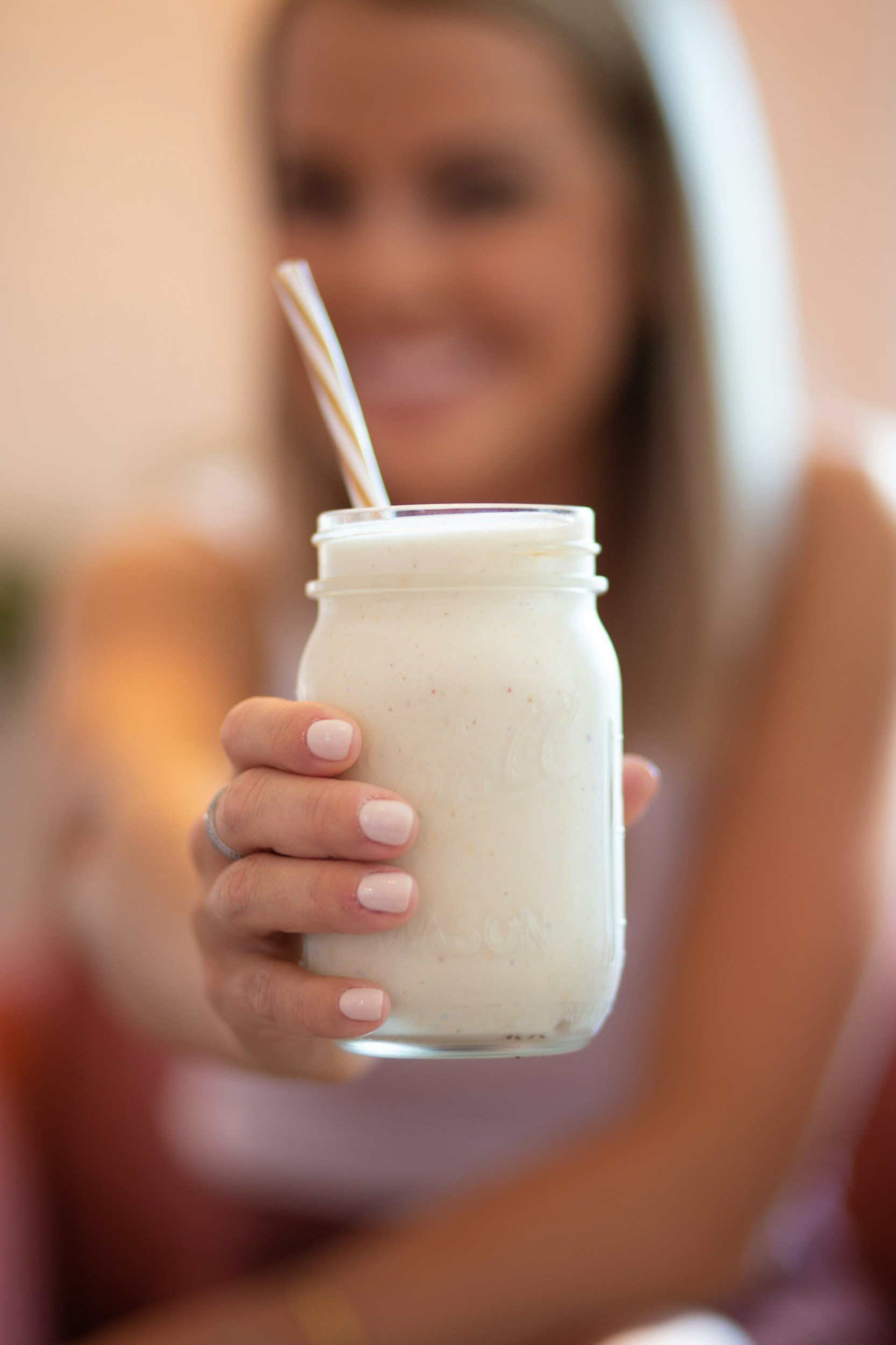A woman with long brown hair and light skin holding a glass jar of creamy white beverage with a spoon inside, smiling in the background.