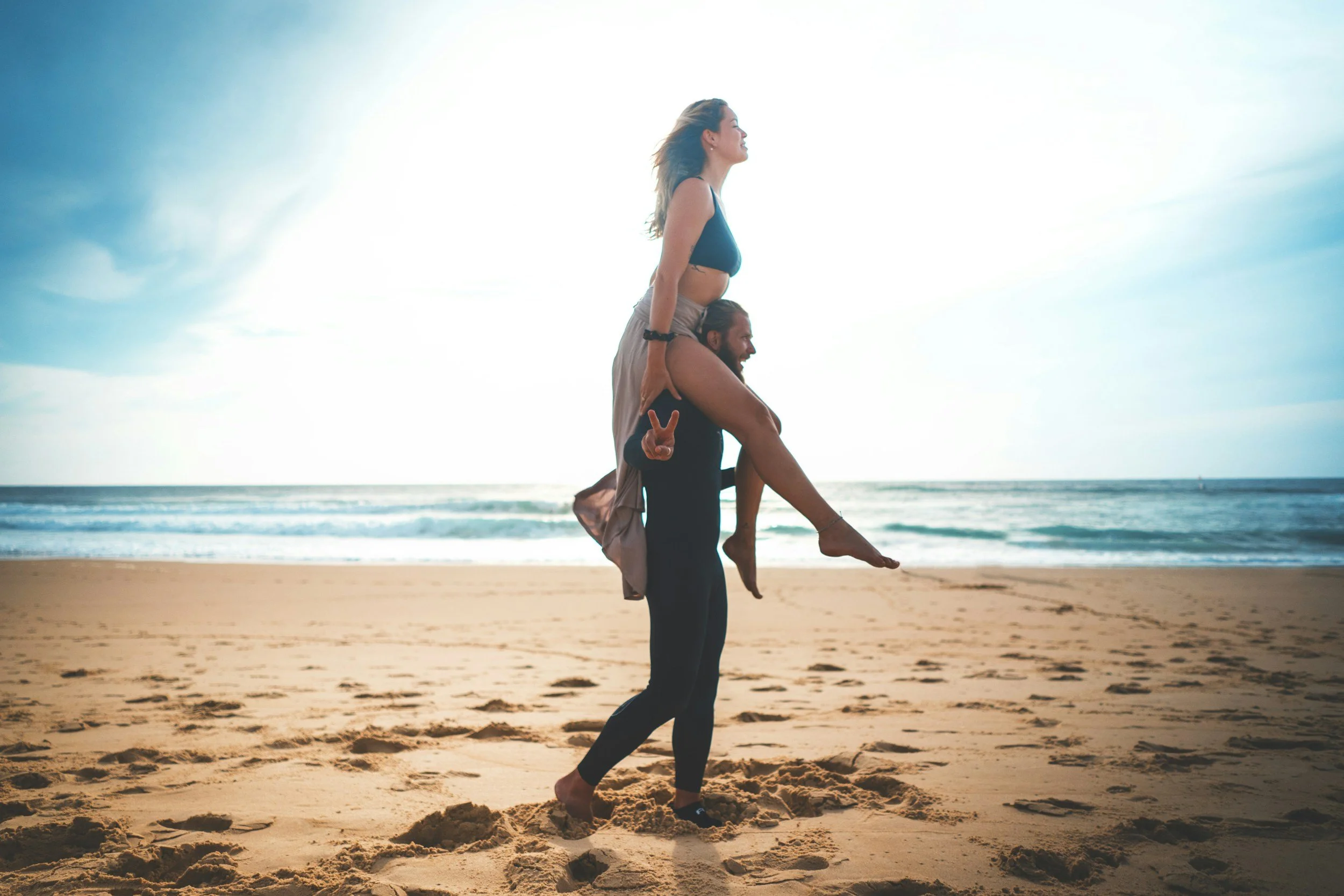 Two people at the beach, with one person giving the other a piggyback ride, both smiling and enjoying the moment, with the ocean and blue sky in the background.