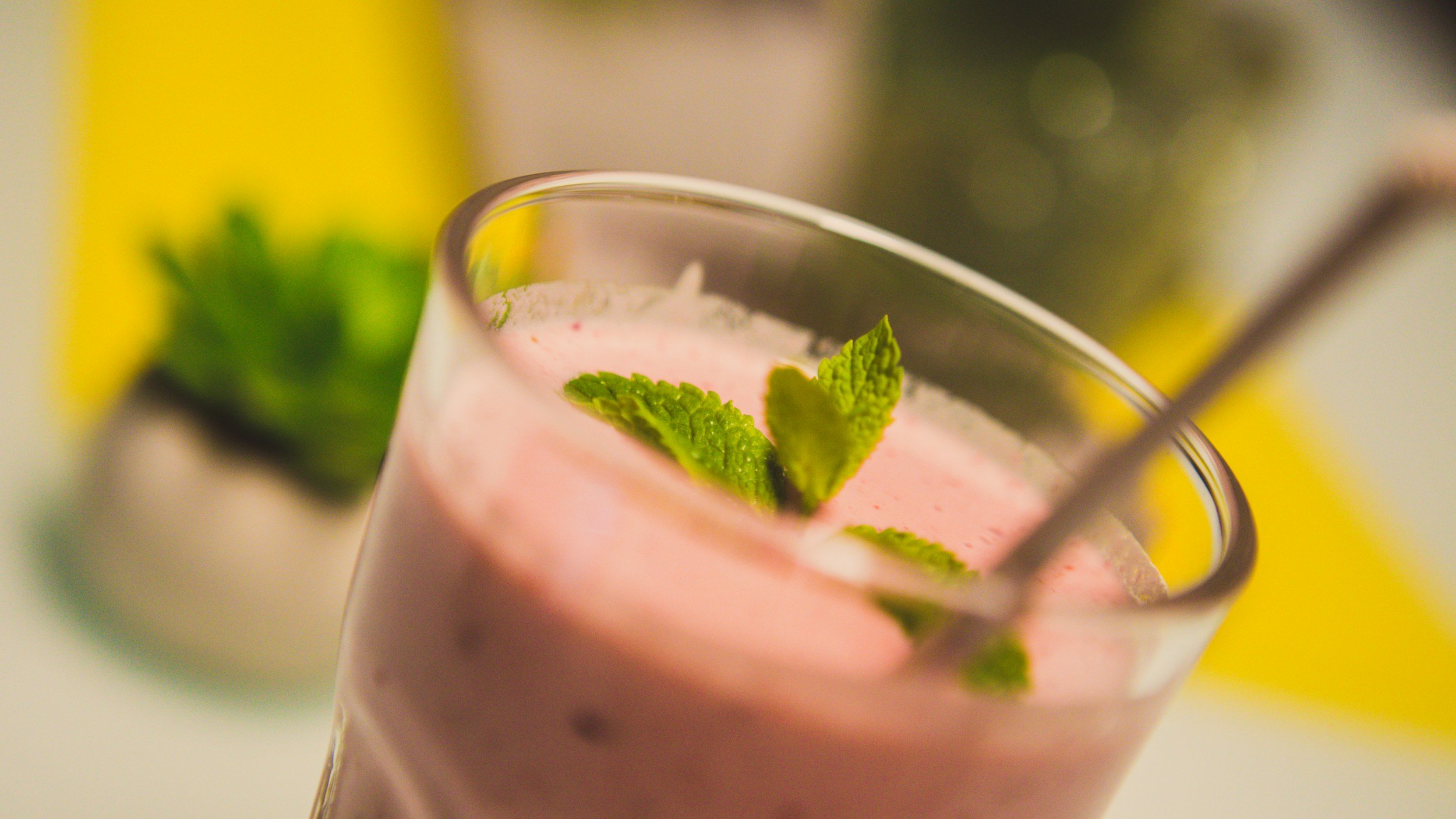 A close-up of a pink frothy drink in a glass garnished with fresh mint leaves, with a blurred yellow background.