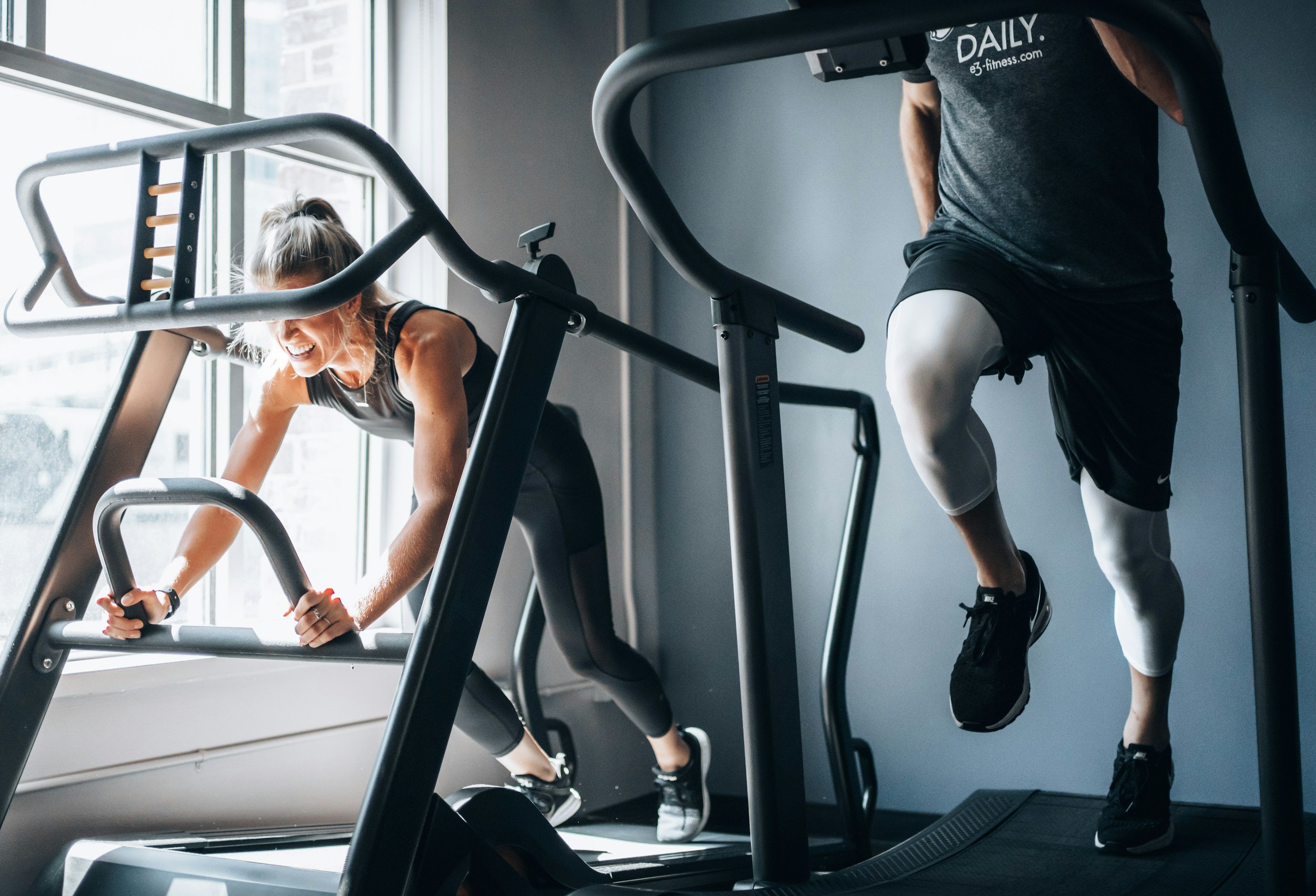 Two people working out on a treadmill at the gym, with natural light coming through large windows.