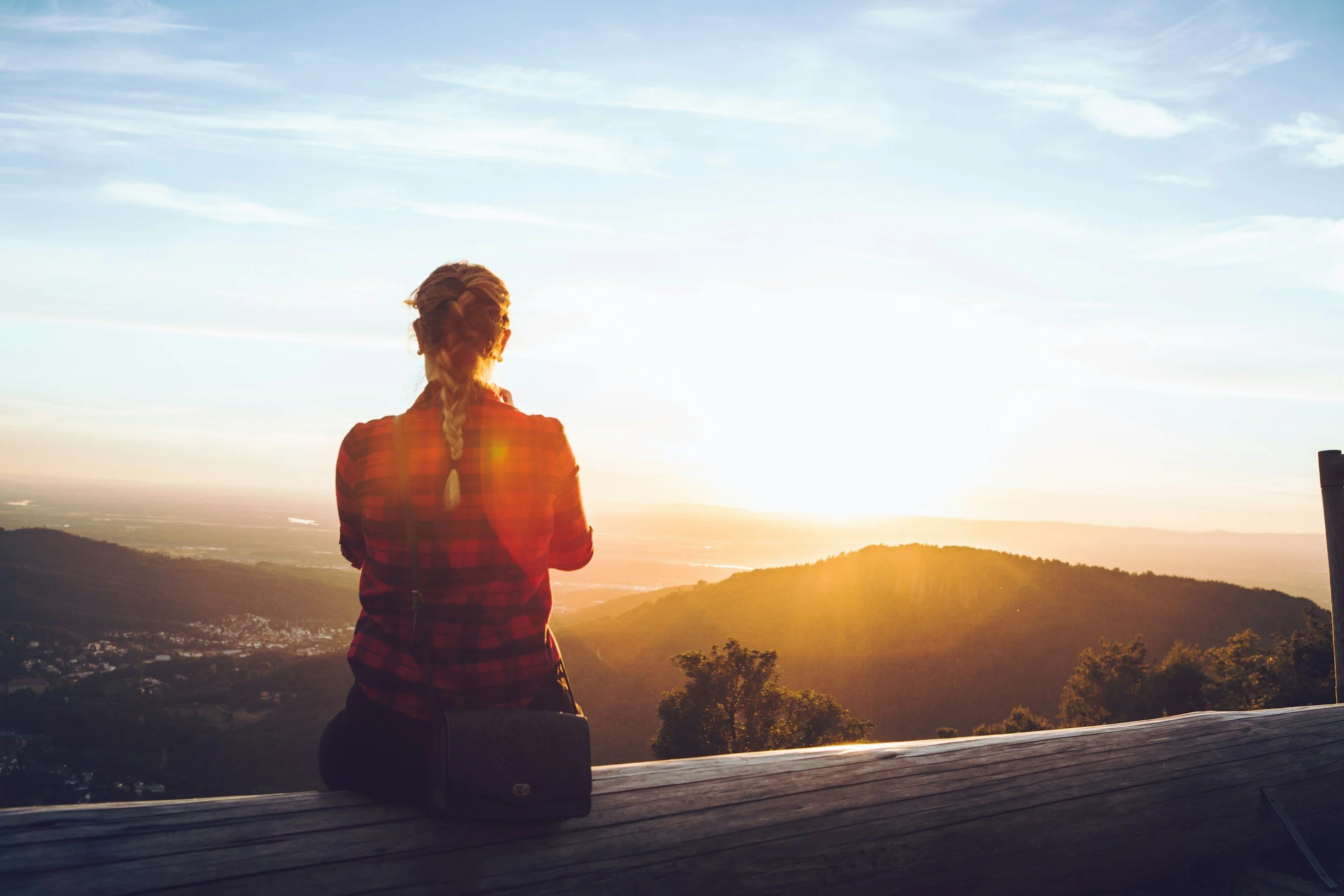 A woman with braided hair in a red plaid shirt sitting on a wooden surface, watching sunset over mountains and valleys.