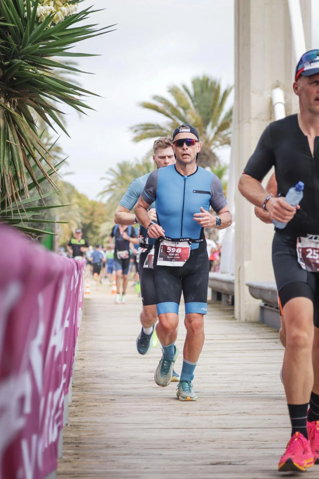 Male runner in a blue and black triathlon outfit, wearing sunglasses and a cap, running on a wooden pathway during a race, with palm trees in the background and other participants in the background.