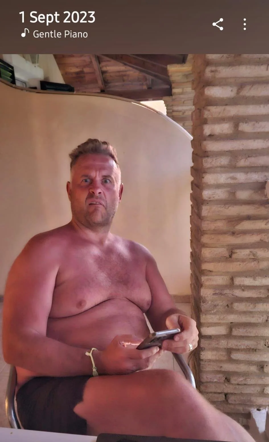 A shirtless man with a surprised or confused expression is sitting indoors, holding a smartphone in his right hand. The background features a brick wall and wooden ceiling with exposed beams.
