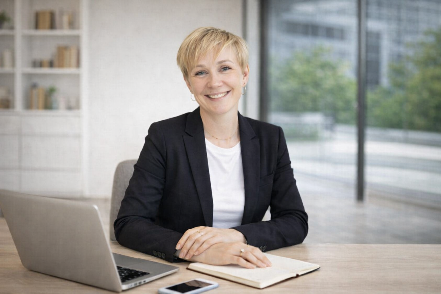 Femme d'affaires souriante assise à un bureau avec un ordinateur portable, un carnet et un smartphone, dans une salle moderne avec grandes fenêtres et bibliothèque.