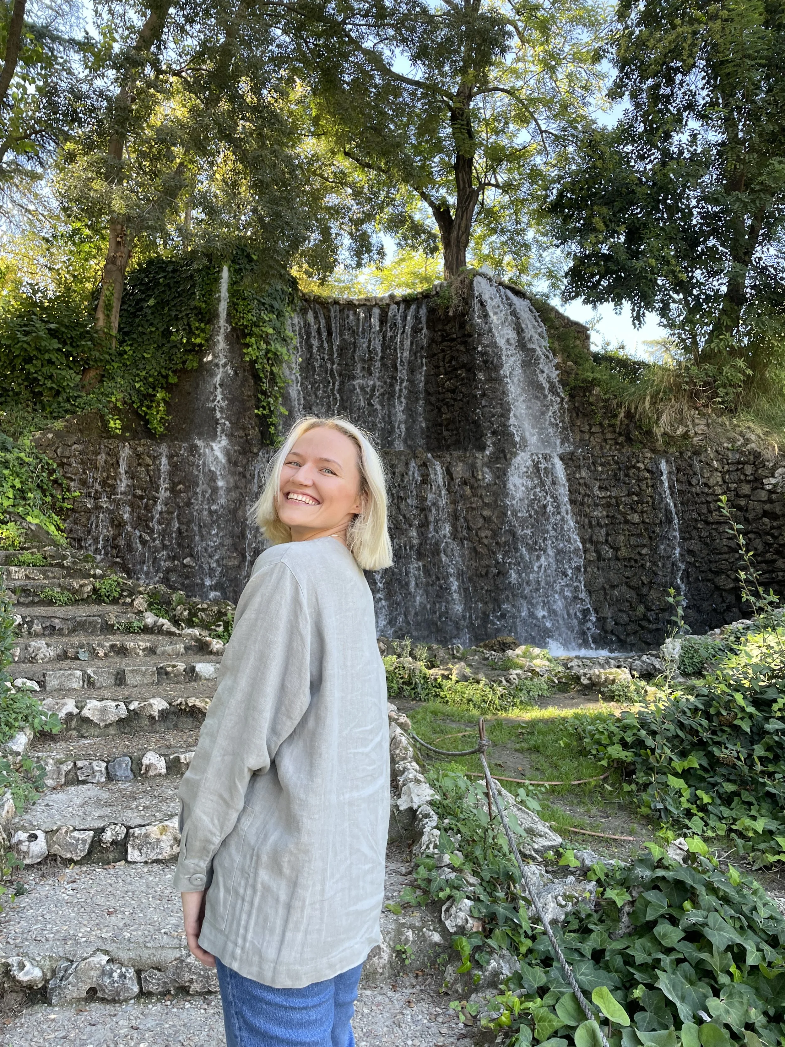 A woman smiling and standing in front of a cascading waterfall with green foliage and trees surrounding it.