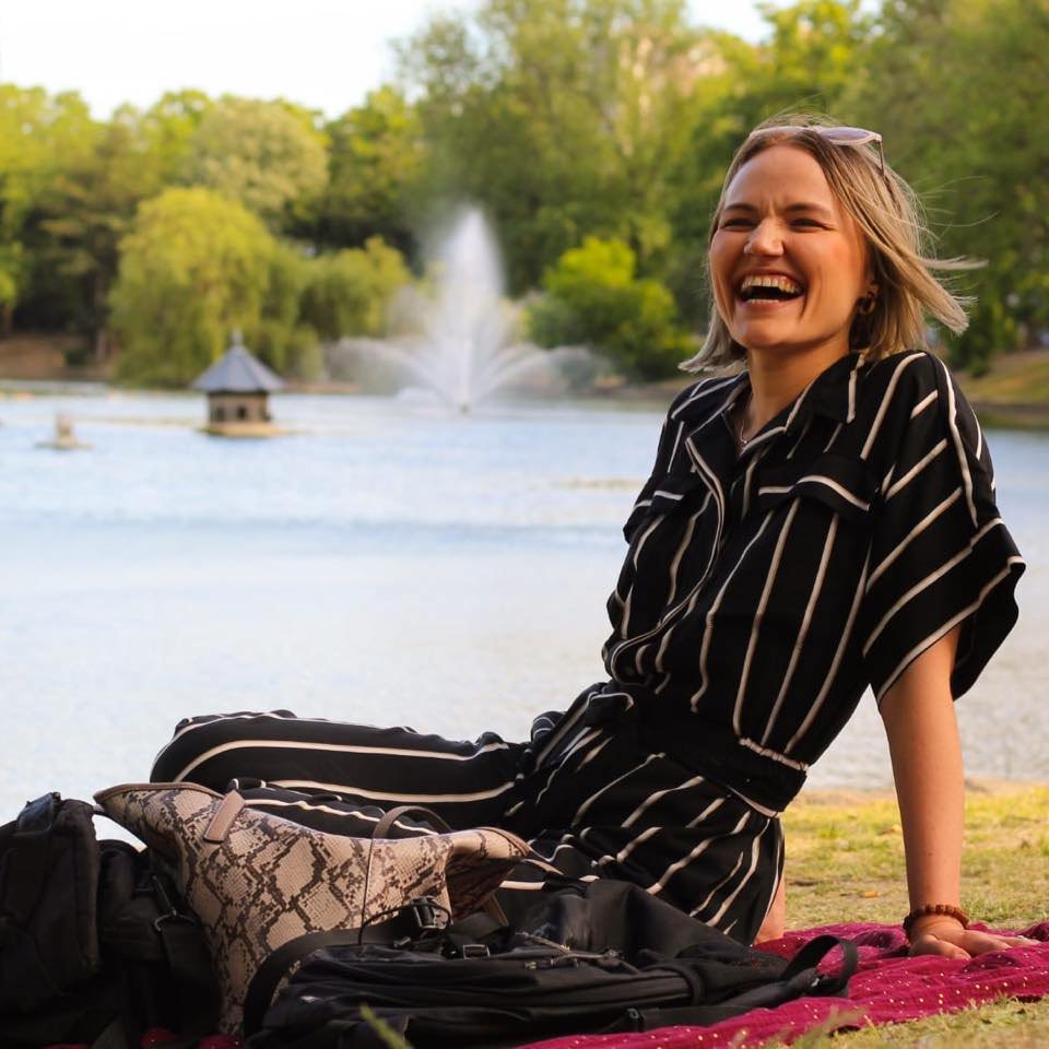 A woman sitting on the ground near a lake, smiling and laughing, with a fountain and trees in the background.