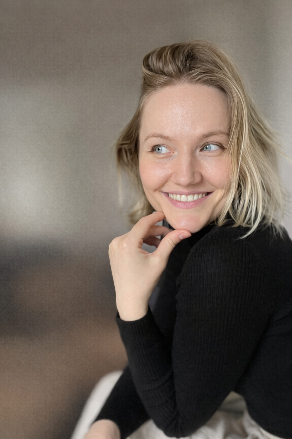 A young woman with blonde hair and blue eyes, smiling and looking to the side, wearing a black sweater.