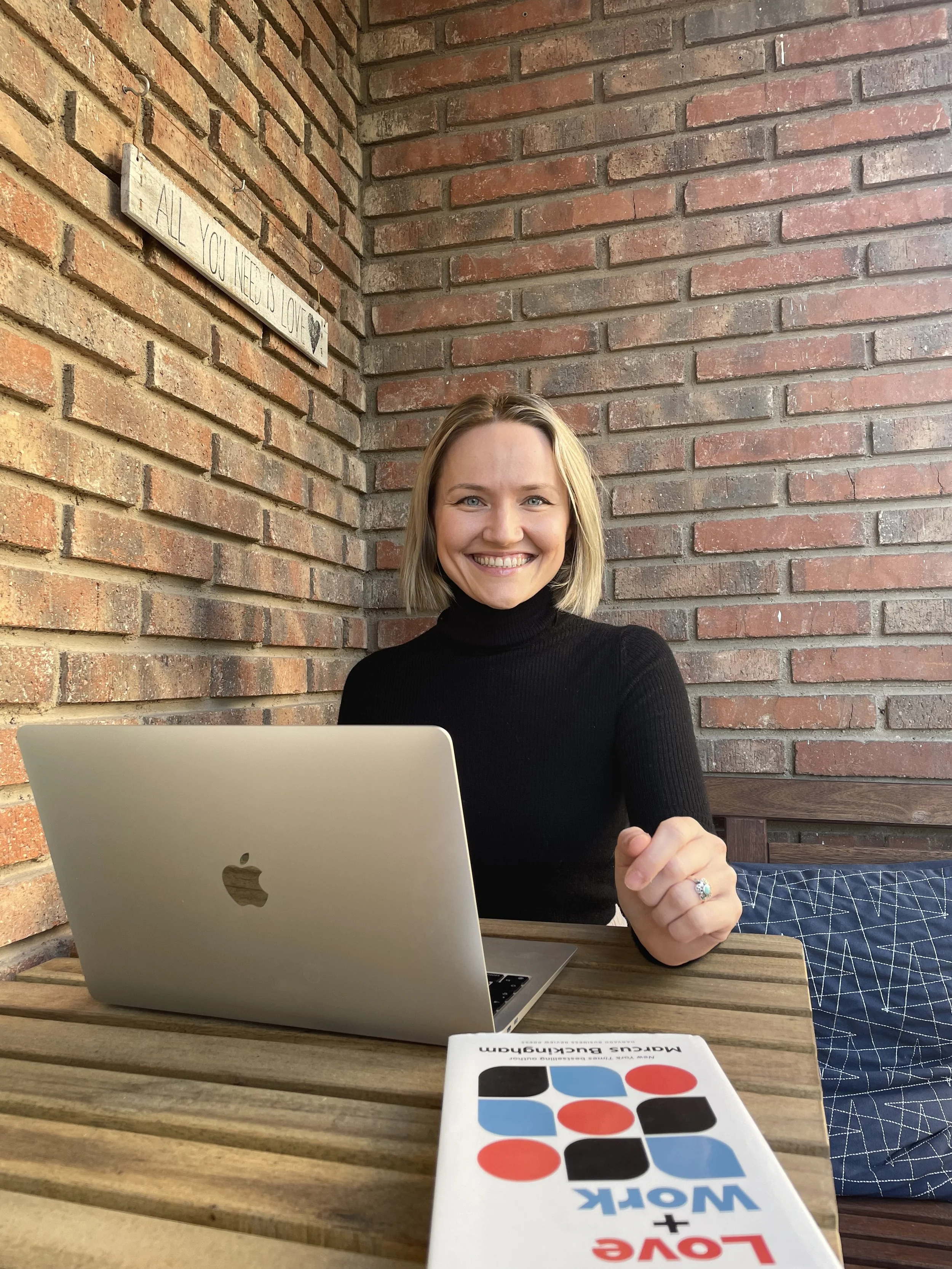 A smiling woman with shoulder-length blonde hair, wearing a black turtleneck, sitting at a wooden table with a silver MacBook laptop in front of her. The background features a brick wall with a decorative sign that reads, 'All you need is love.' A brochure or booklet titled 'Love + Work' lies on the table.